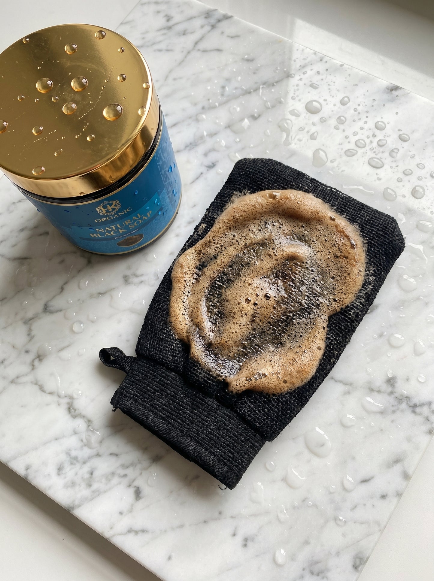 Black exfoliating glove with soap on a marble surface next to a blue container with a gold lid.