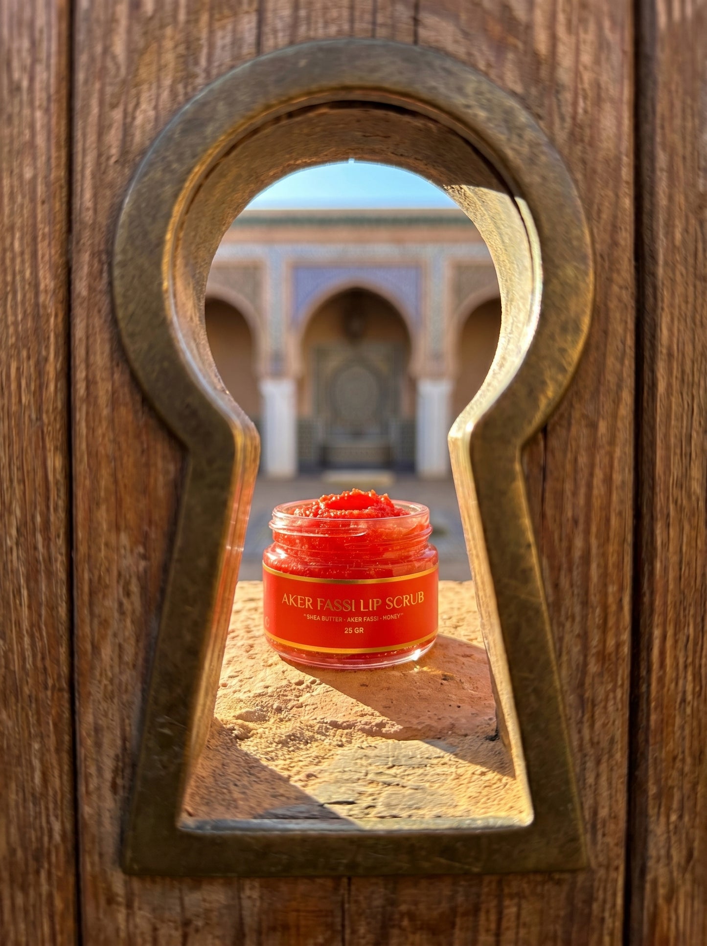 Red jar of lip scrub through a wooden keyhole with a blurred architectural background