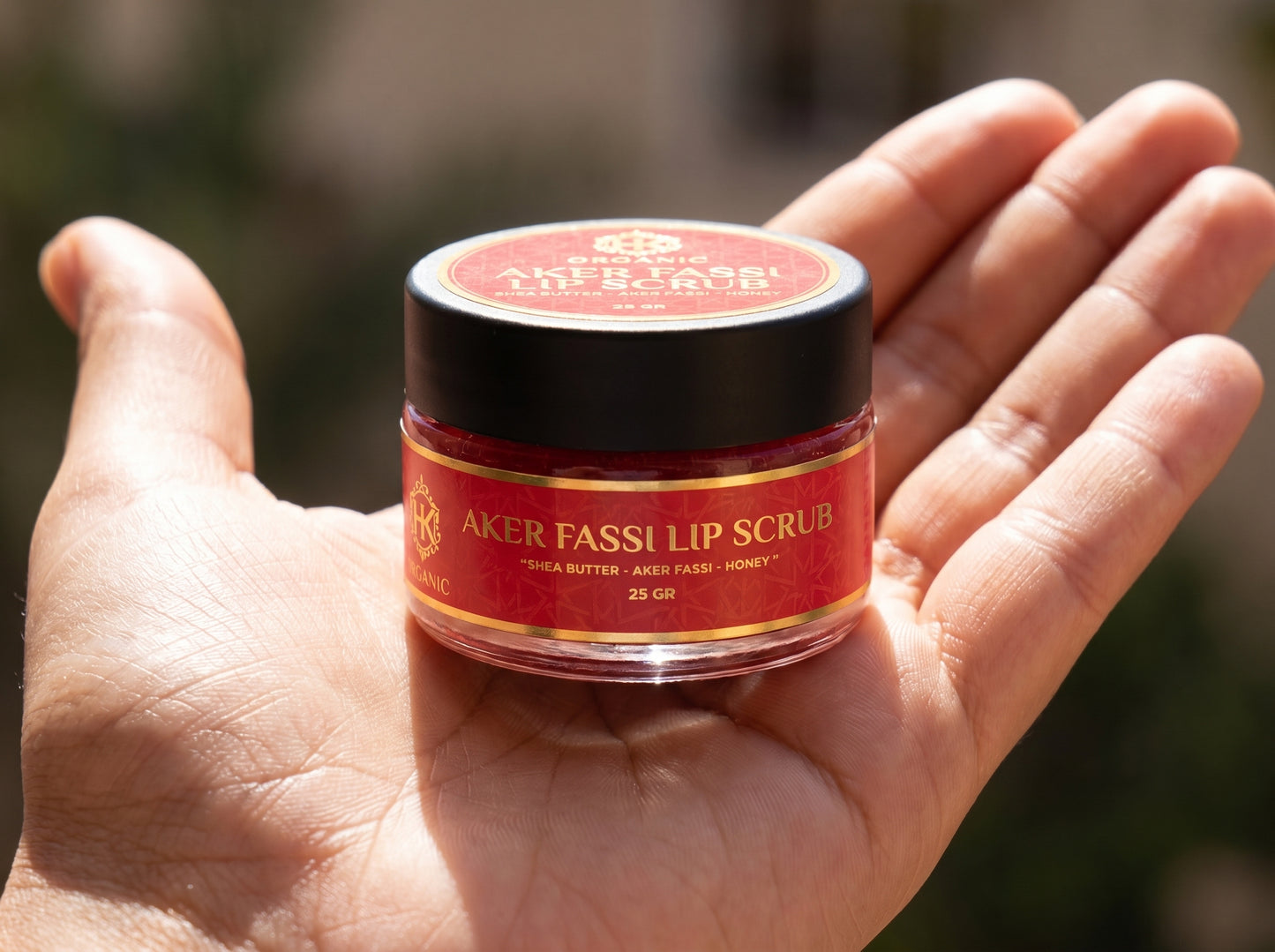 Hand holding a jar of Aker Fassi lip scrub with a blurred background