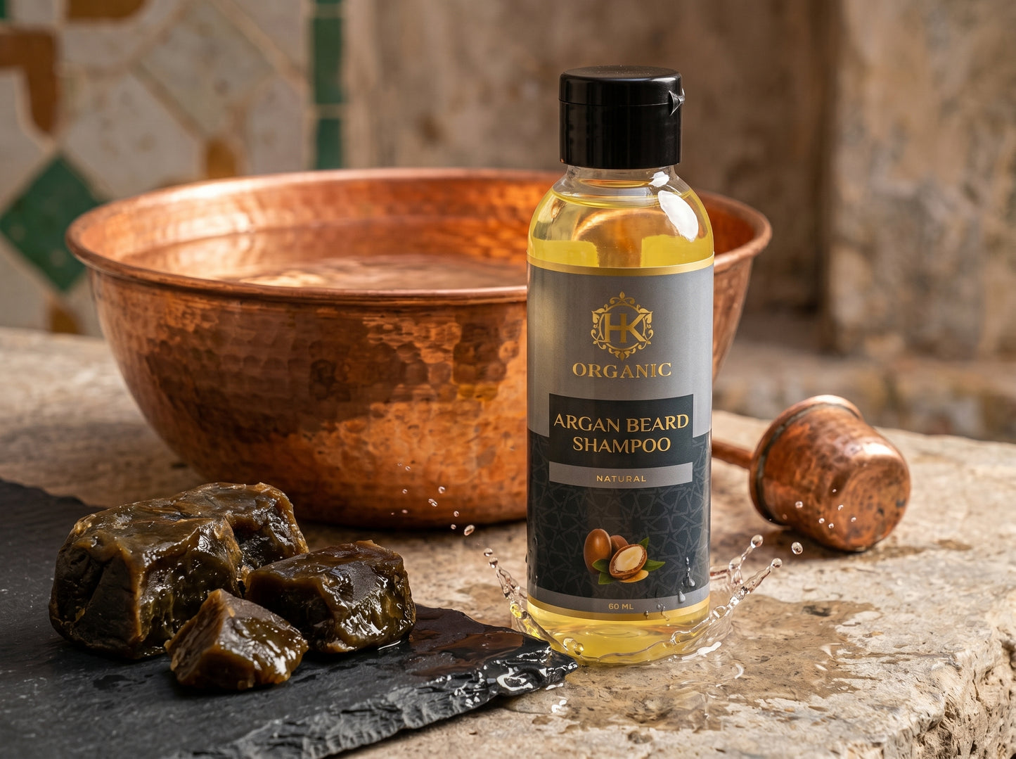 Bottle of Organic Argan Beard Shampoo with argan stones and a copper bowl on a stone surface.