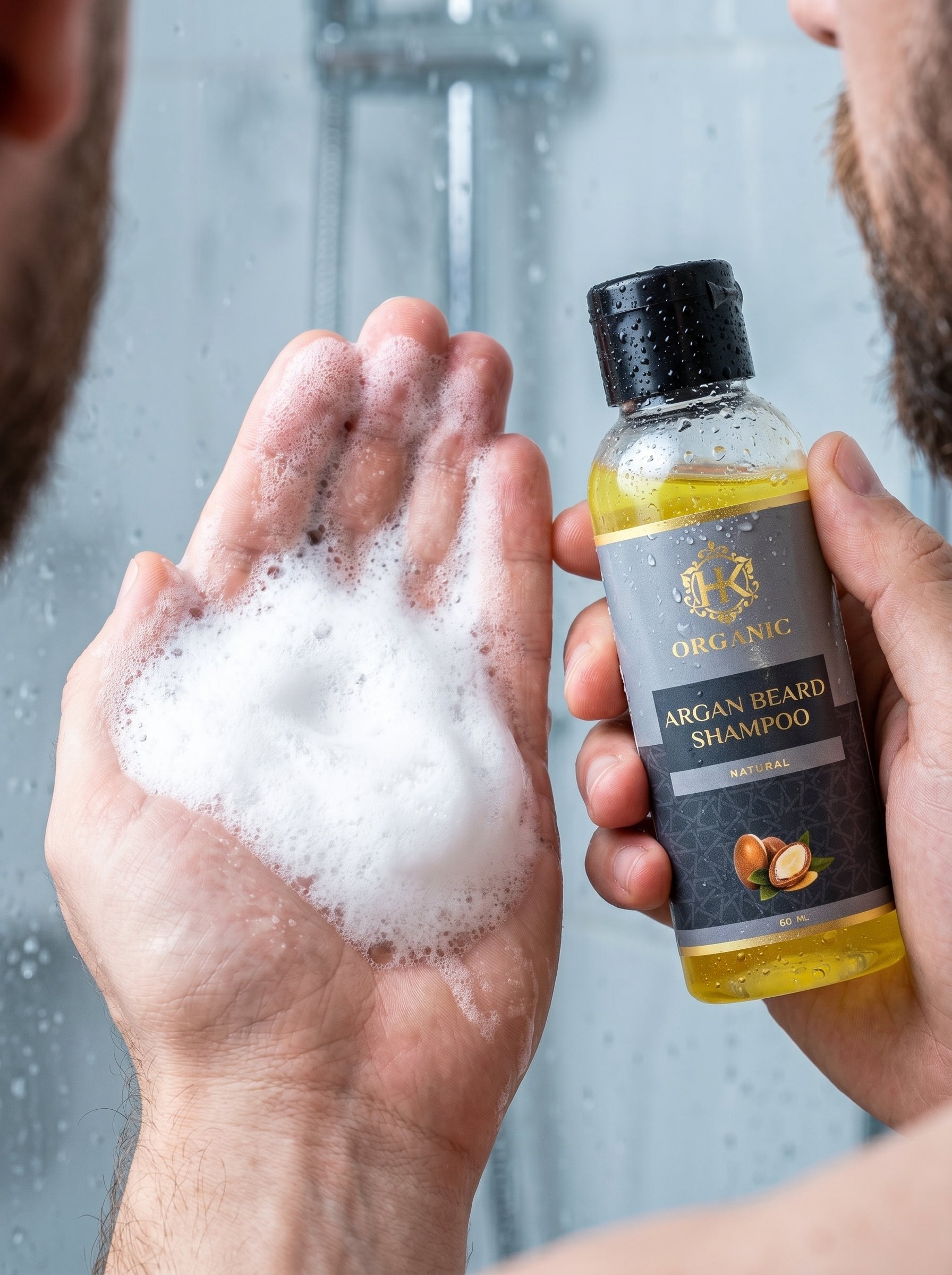 Person holding a bottle of Organic Argan Beard Shampoo with soapy hands in a shower setting.