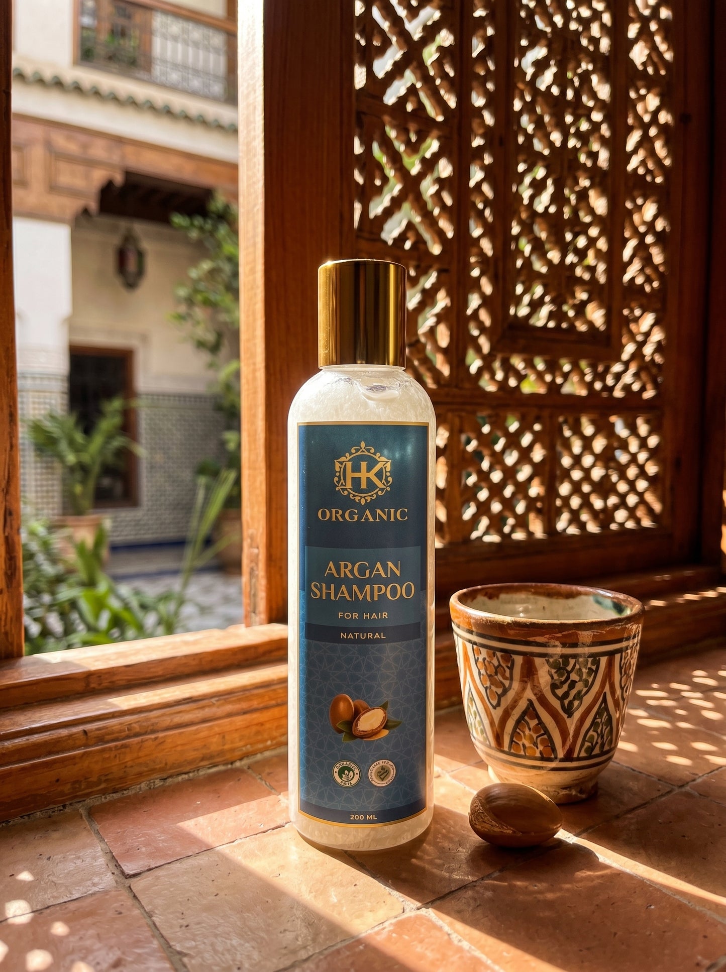 Bottle of organic argan shampoo on a sunlit windowsill with a decorative pot and wooden window.