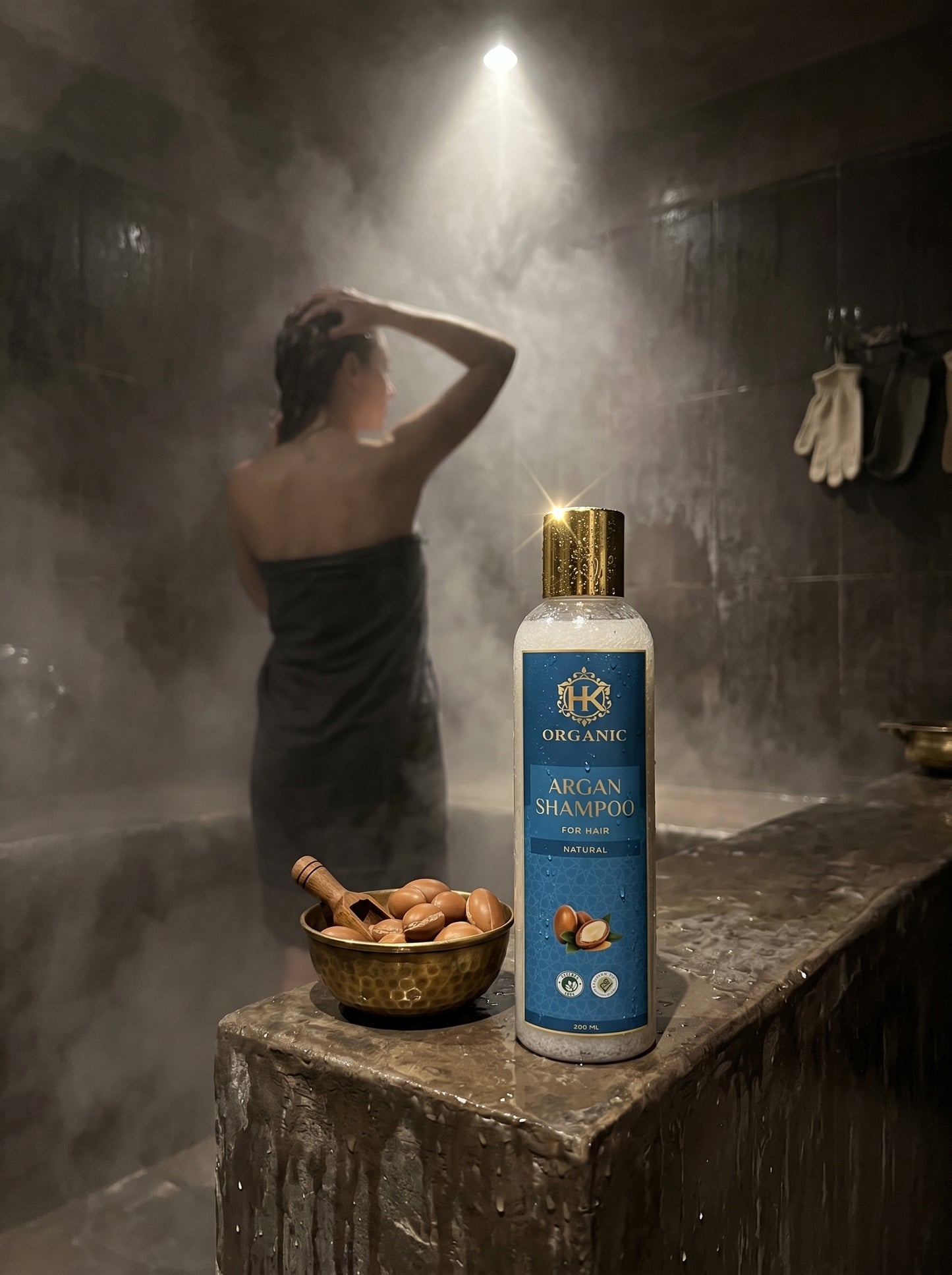 Bottle of organic argan shampoo on a stone surface with a person showering in the background.