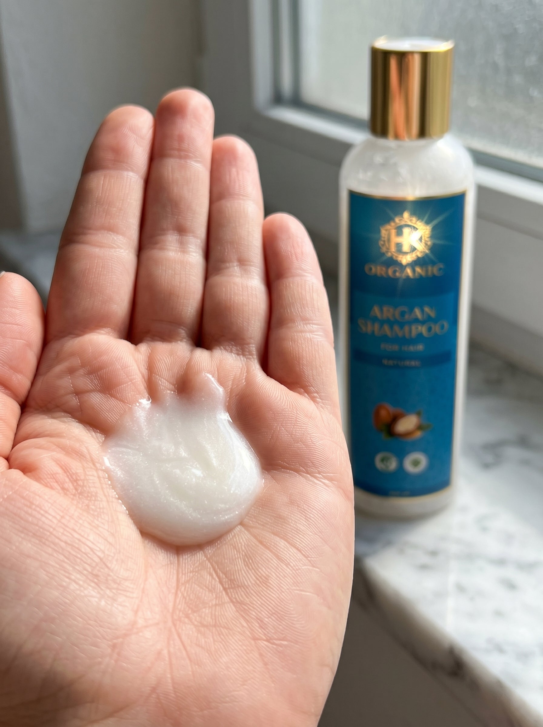 Hand holding a small amount of shampoo next to a bottle of shampoo on a surface.
