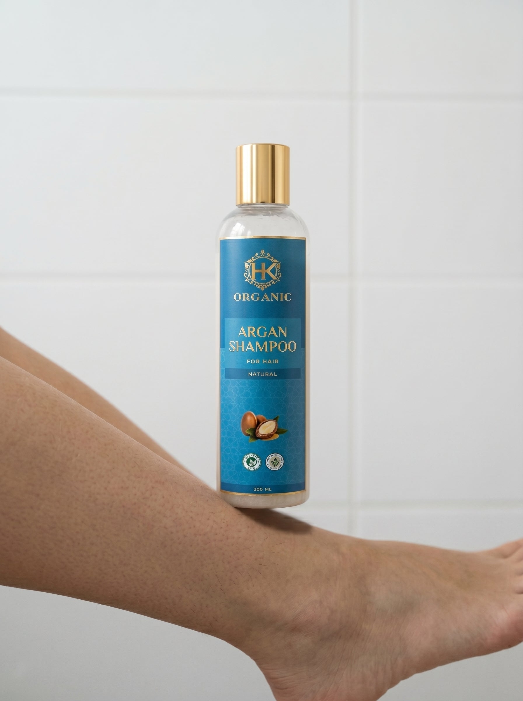 Bottle of organic argan shampoo held by a person against a white tiled wall.
