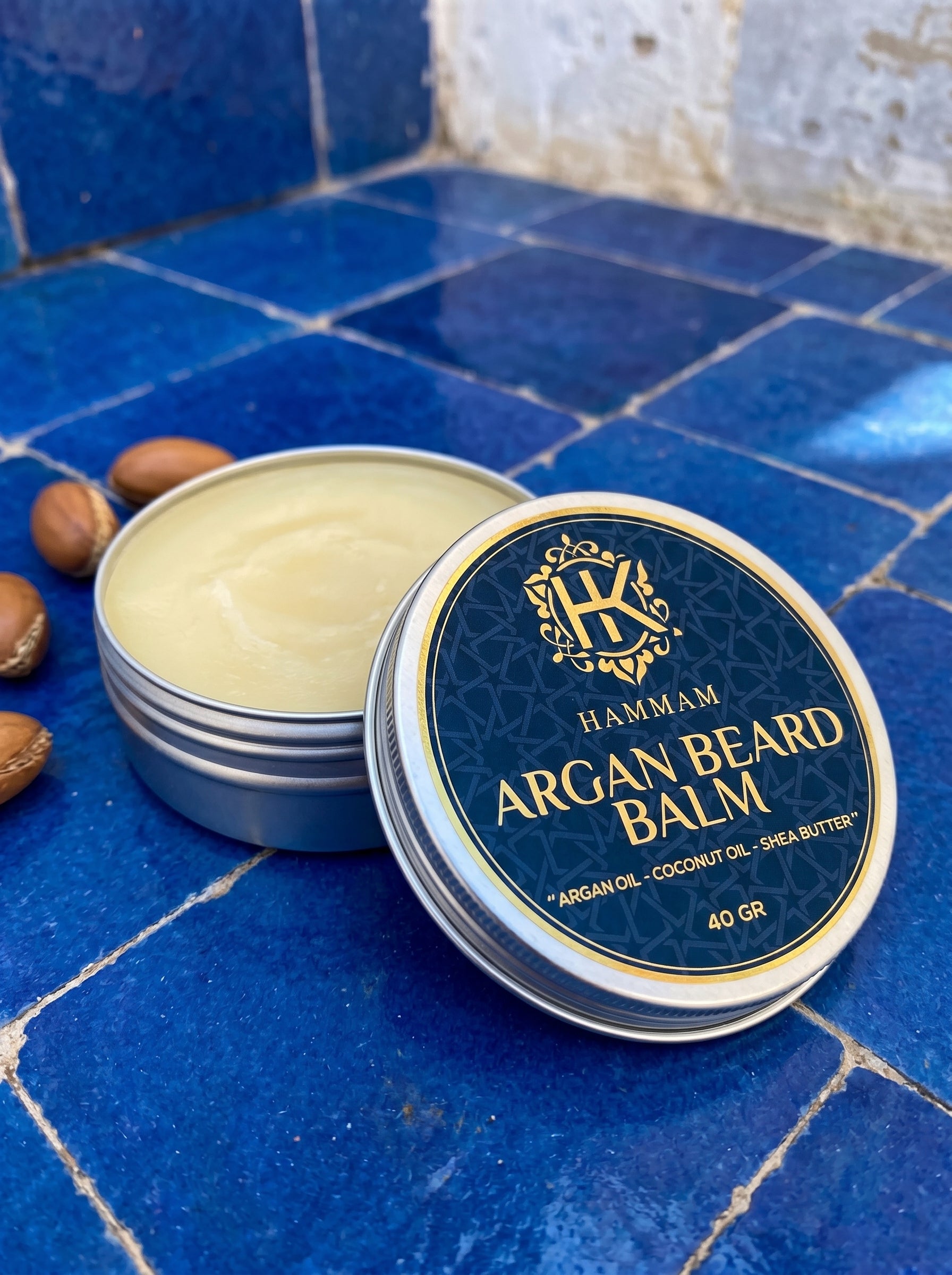 Hammam Argan Beard Balm container on a blue tiled surface with argan seeds.