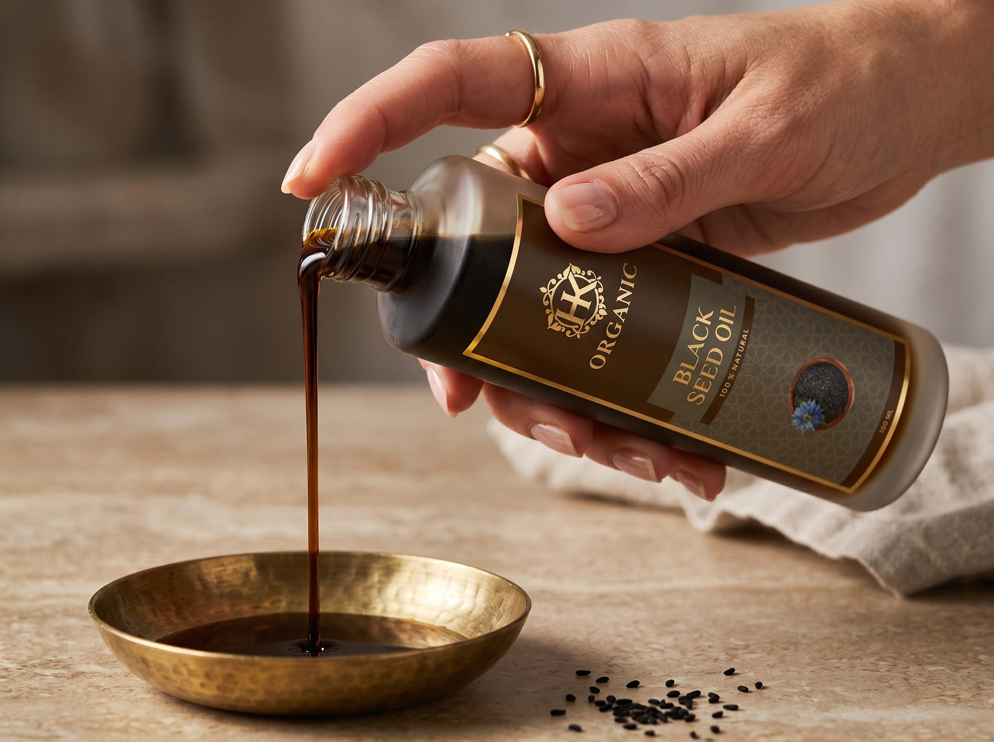 Hand pouring black seed oil from a bottle labeled 'Organic Black Seed Oil' into a brass bowl on a wooden surface.