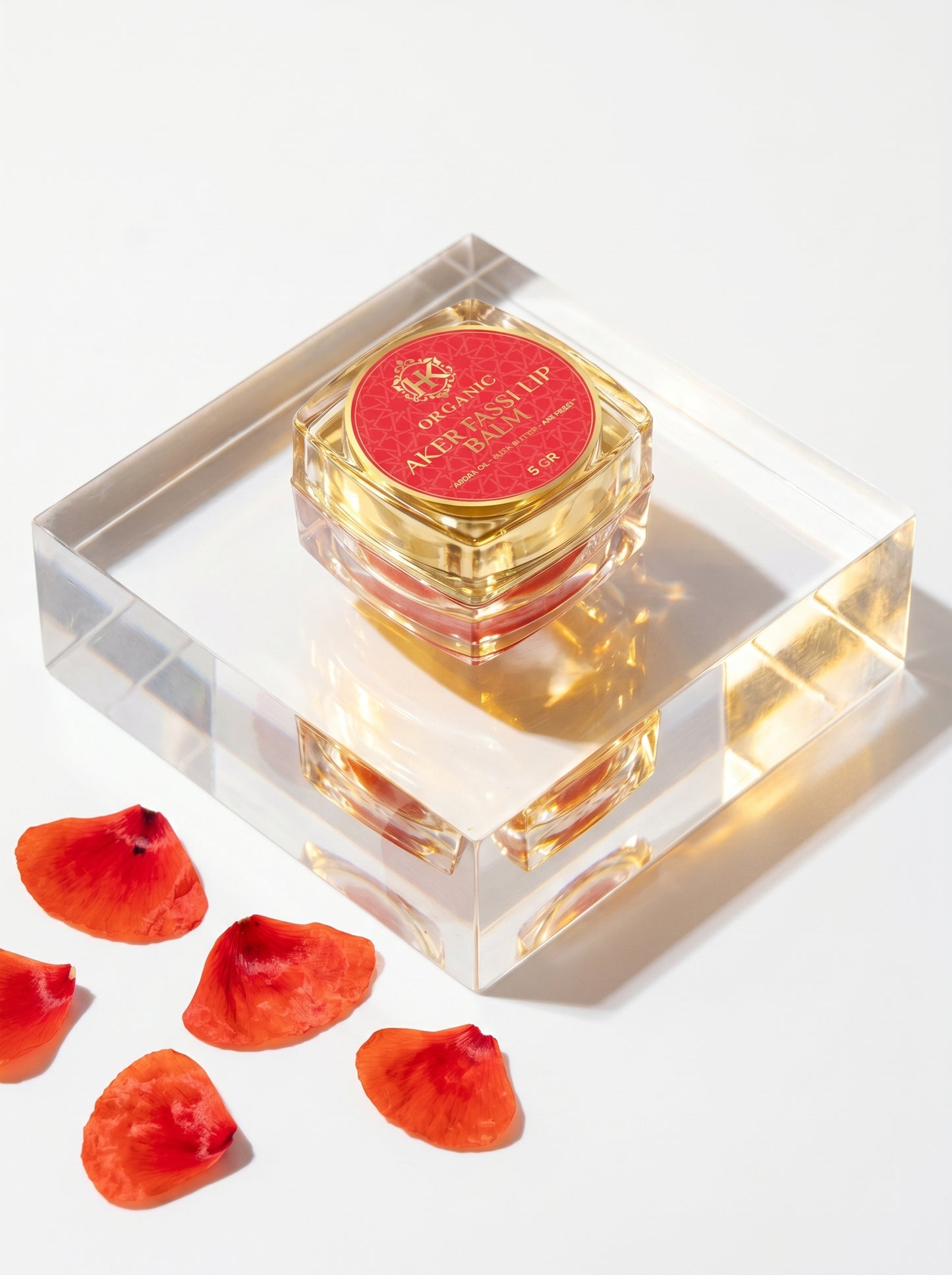 Perfume bottle with red cap on a clear stand, surrounded by red petals on a white background