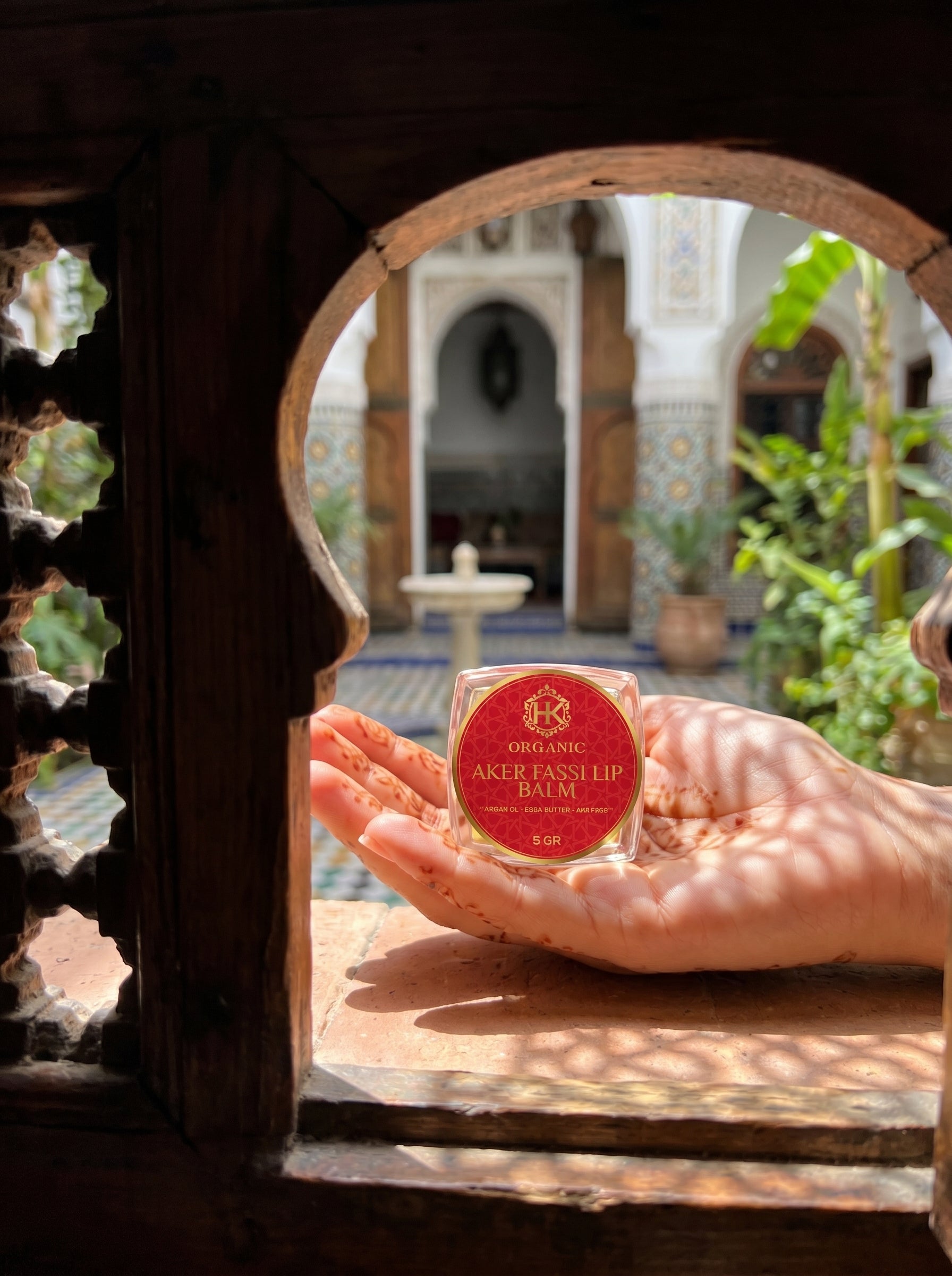 Hand holding a jar of 'Aker Fassilh Balm' in front of a decorative window with a garden view.