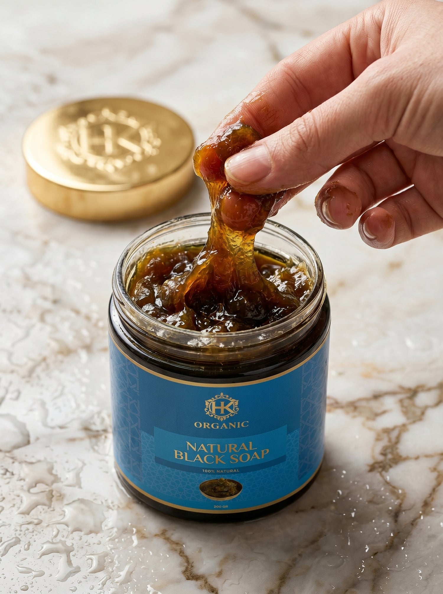 Hand dipping a piece of soap into a jar of natural black soap on a marble surface.