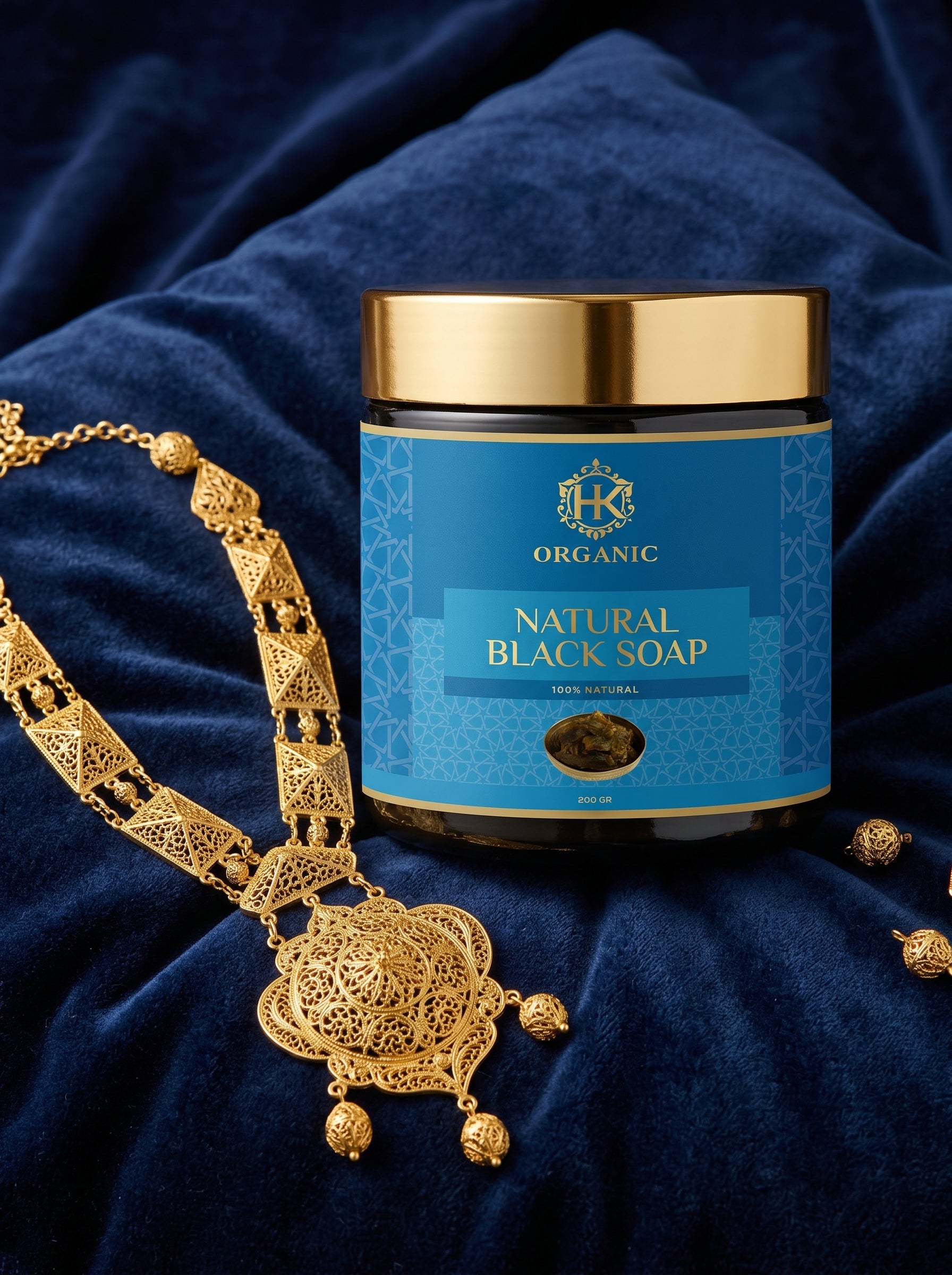 Jar of organic natural black soap with a gold lid on a blue fabric background, next to a gold necklace.