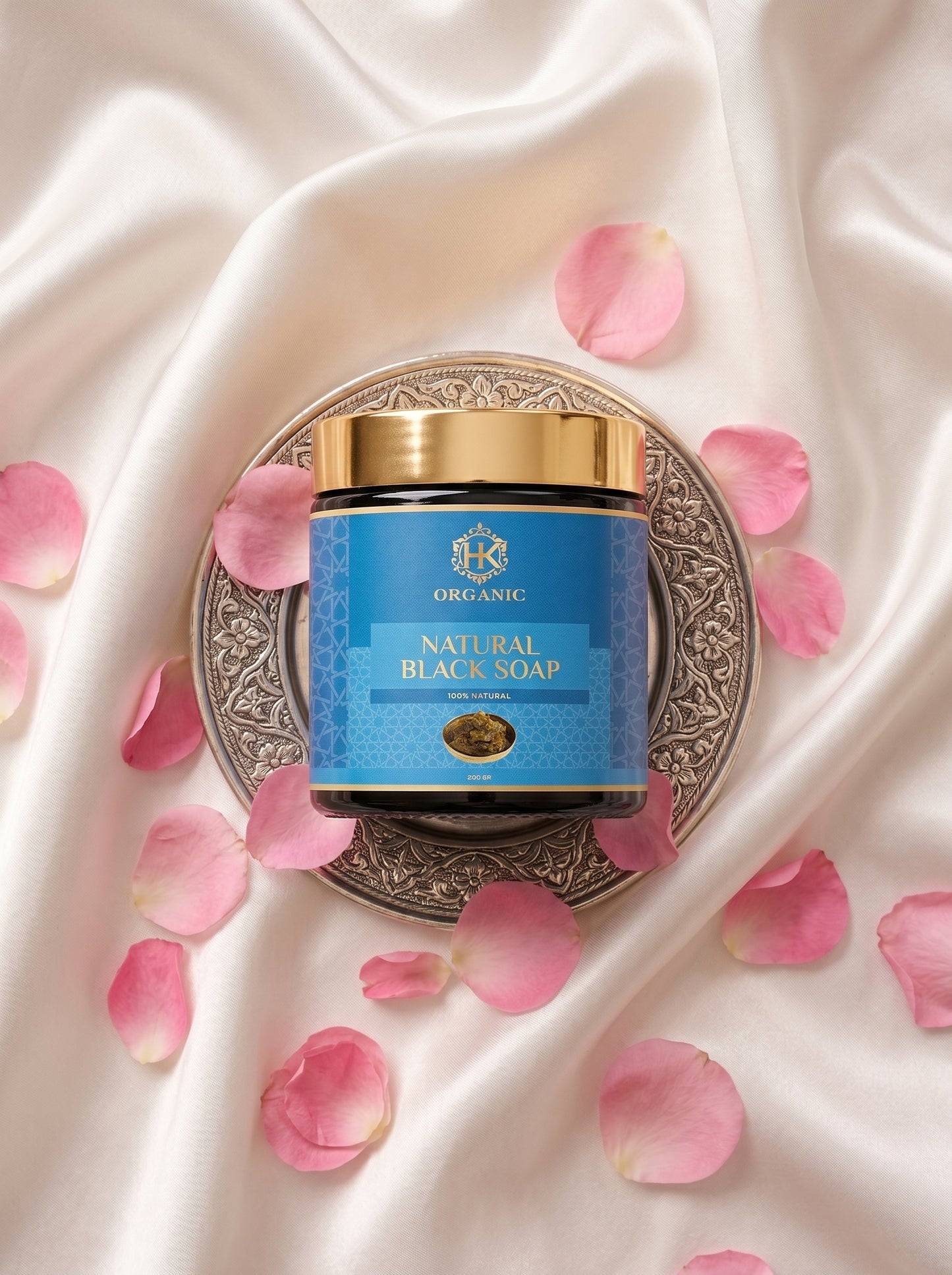 Jar of natural black soap with a gold lid on a decorative plate surrounded by pink rose petals on a satin background.