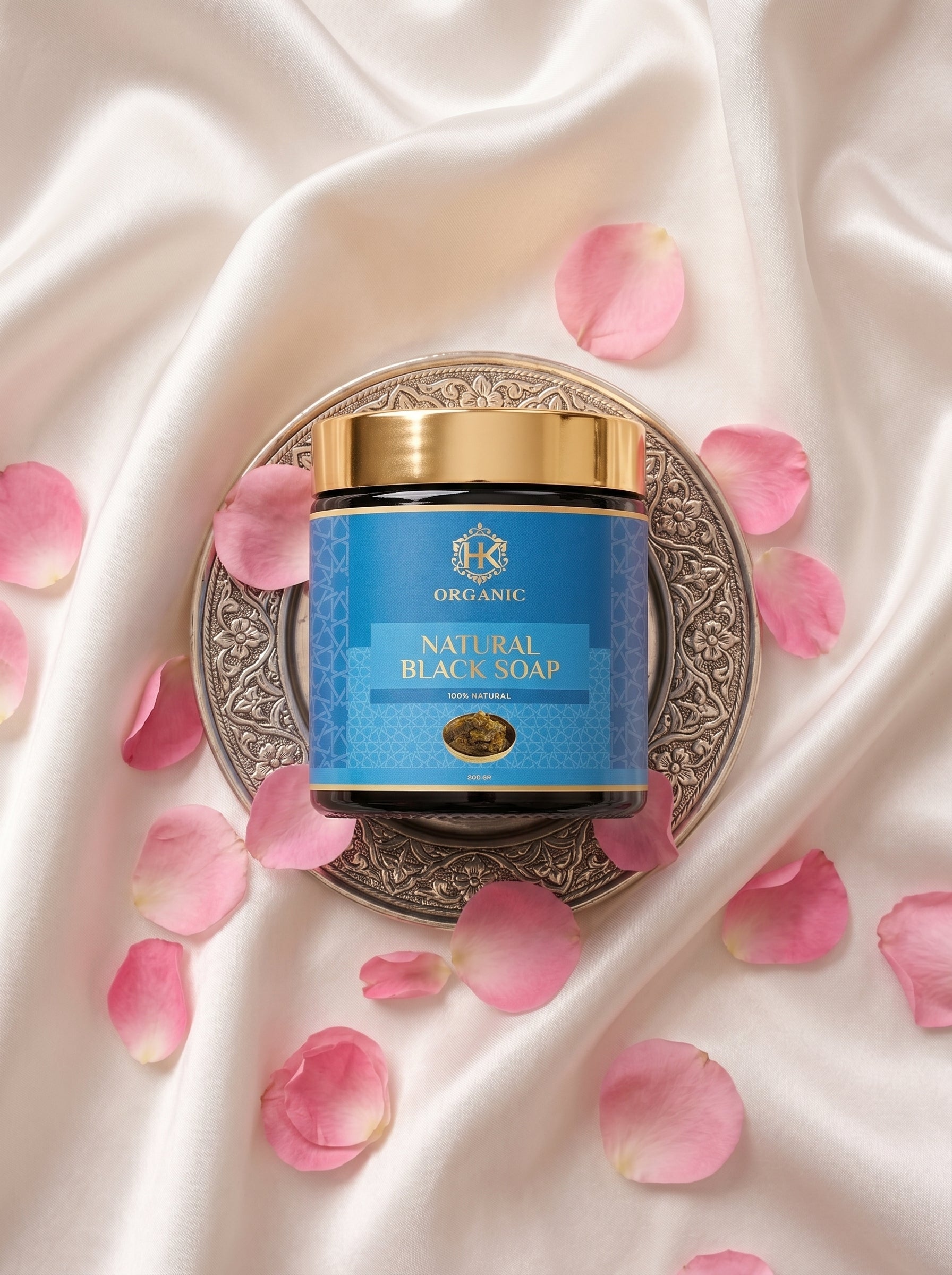 Jar of natural black soap with a gold lid on a decorative plate surrounded by pink rose petals on a satin background.
