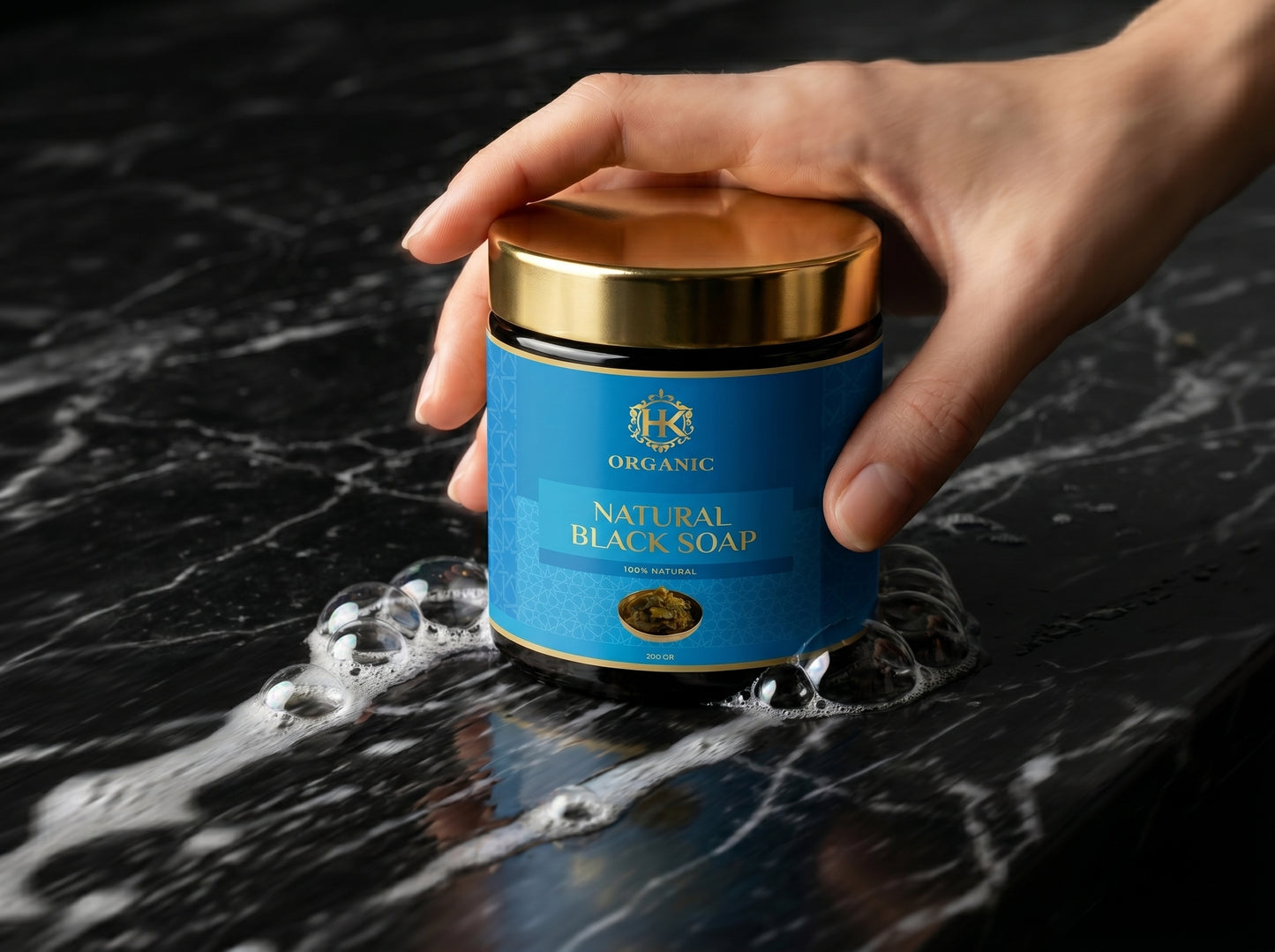 Hand holding a jar of organic natural black soap on a dark surface with water.
