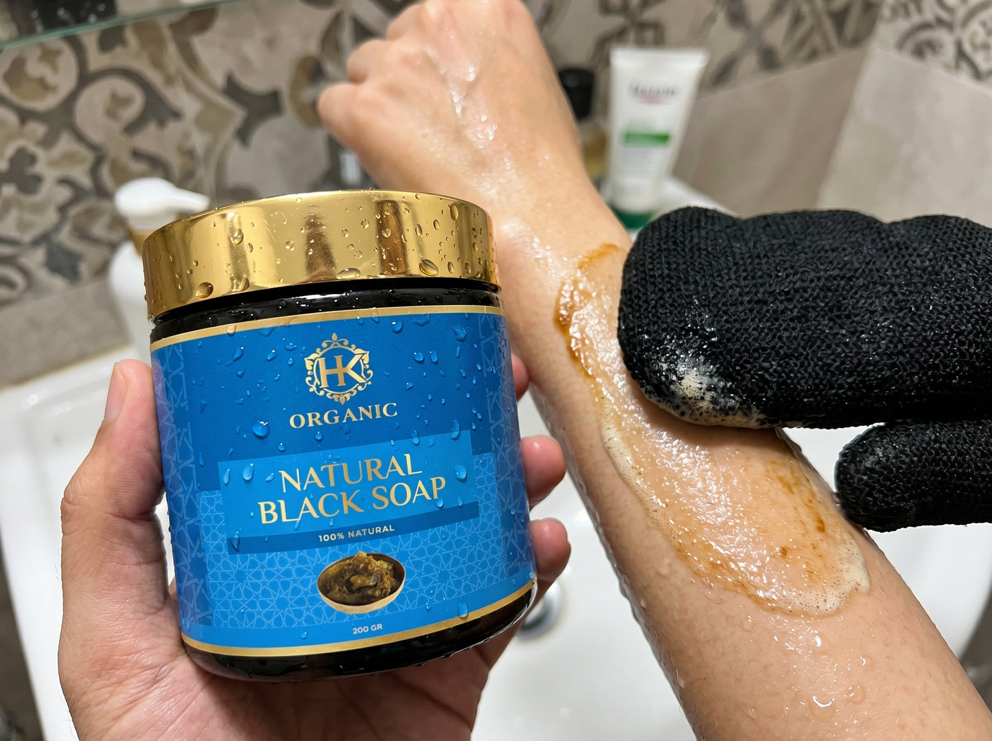 Person applying natural black soap to their hand with a jar of the product held in the other hand.