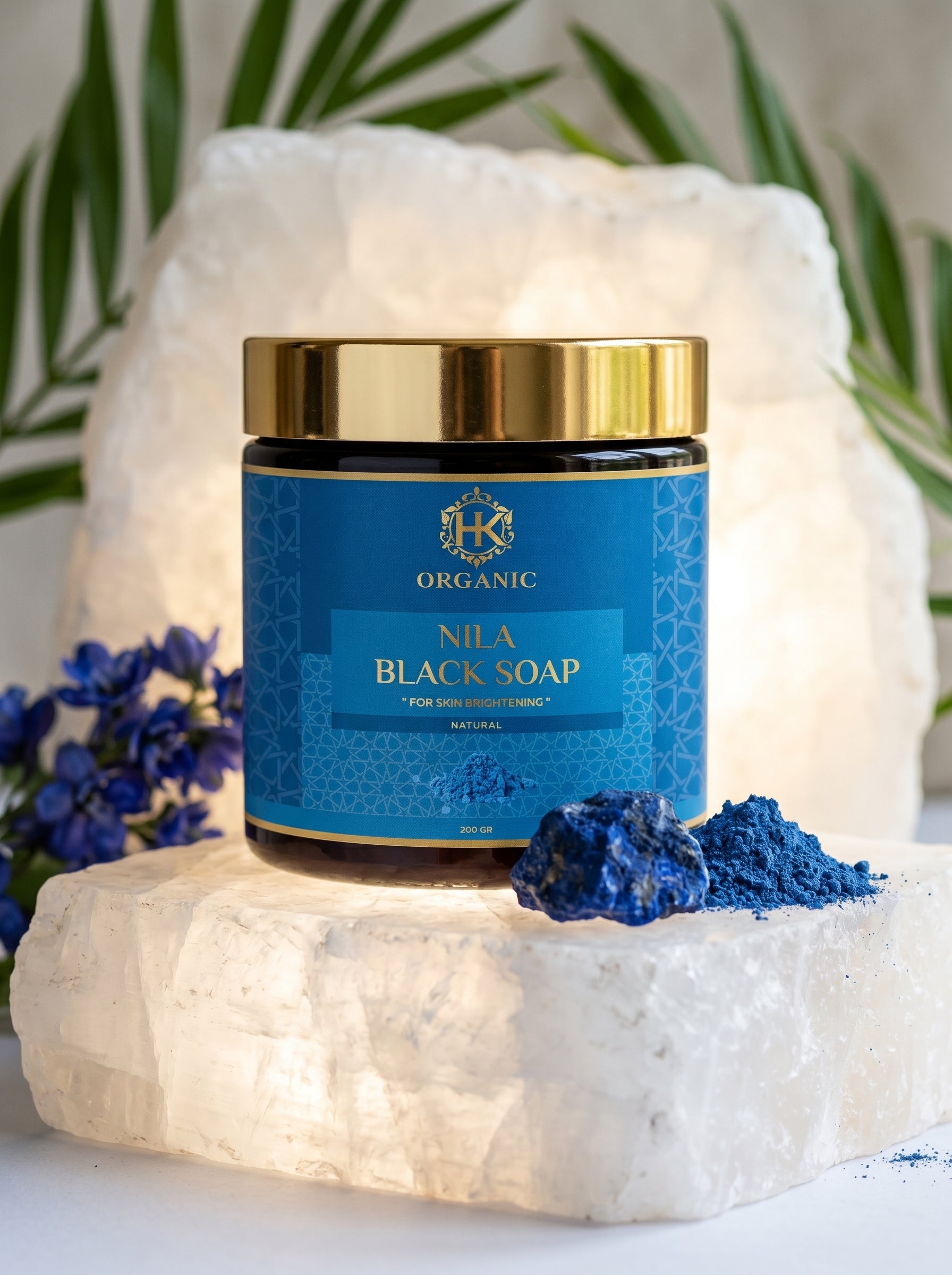 Nila Black Soap jar with gold lid on a white stone surface with blue flowers and green leaves in the background
