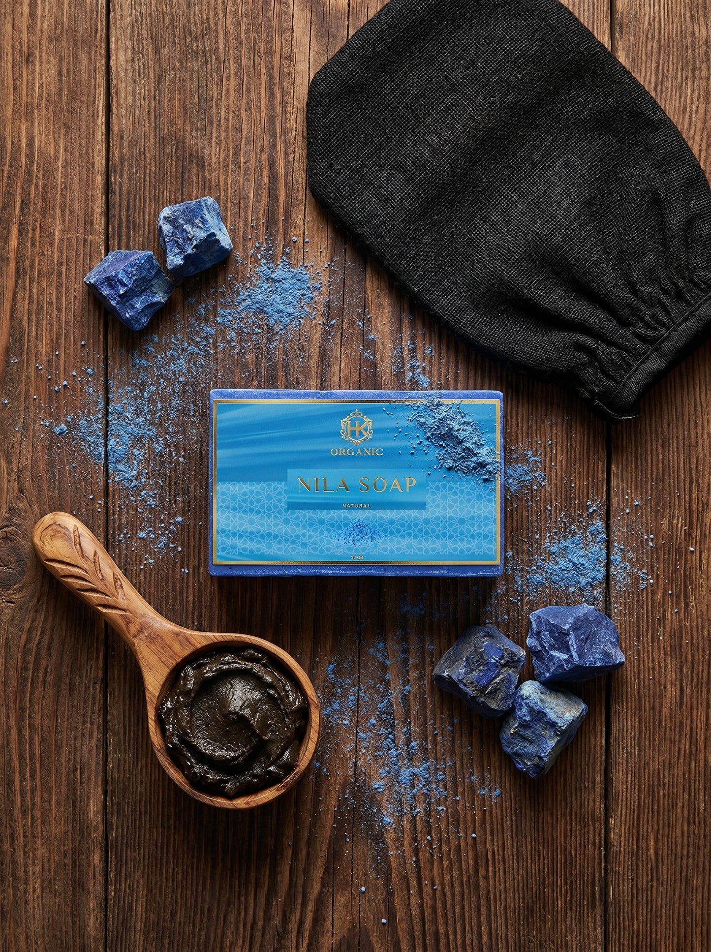 Bar of soap with a wooden spoon containing a dark paste, surrounded by blue lumps and a black glove on a wooden surface.