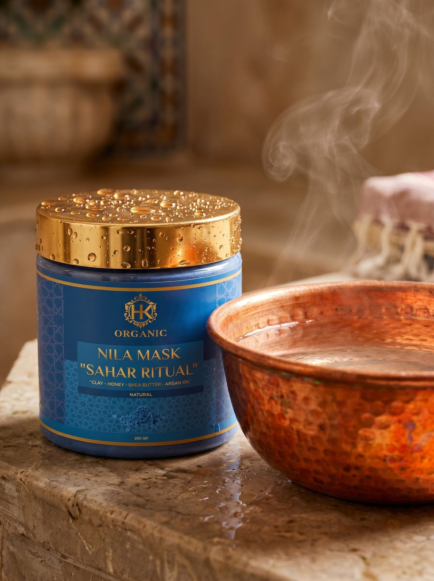 Blue jar of Nila Mask with gold lid next to a copper bowl with steam, on a stone surface.