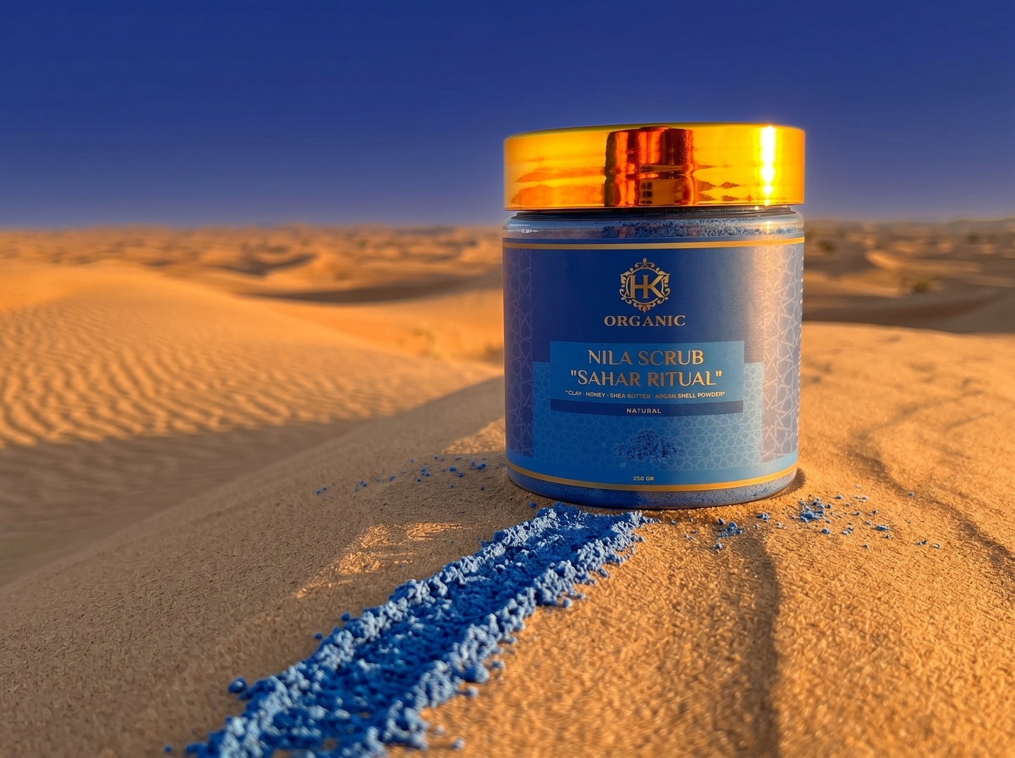 Jar of 'Nila Scrub Sahara Ritual' on sand with blue powder spilling out against a desert backdrop