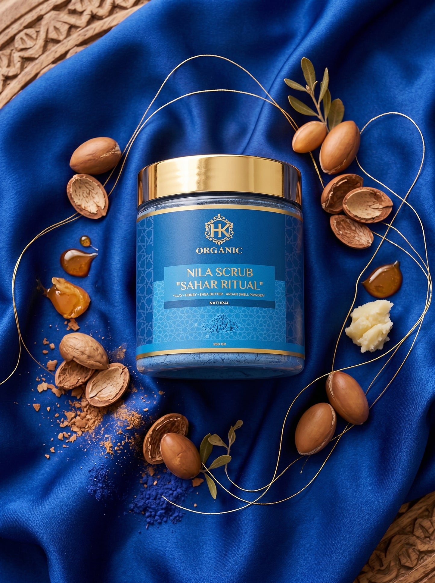 Blue jar of scrub with gold lid on a blue fabric background with nuts and leaves.