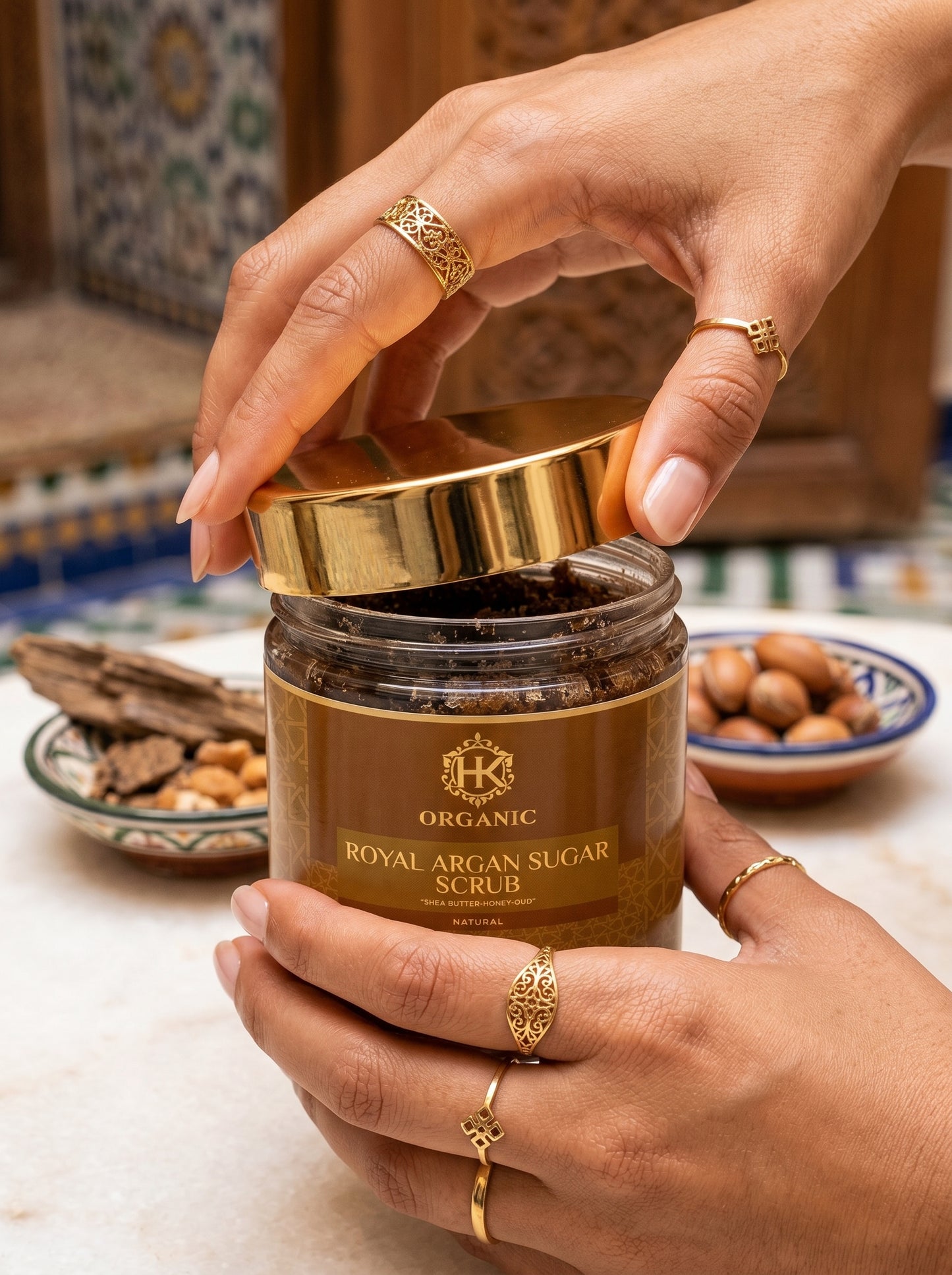 Jar of organic royal argan sugar scrub held by a hand with gold rings on a decorative background.