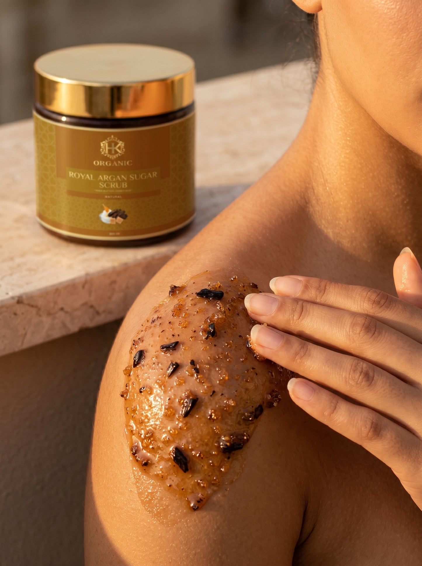 Person applying a brown scrub to their shoulder with a jar of the scrub in the background.