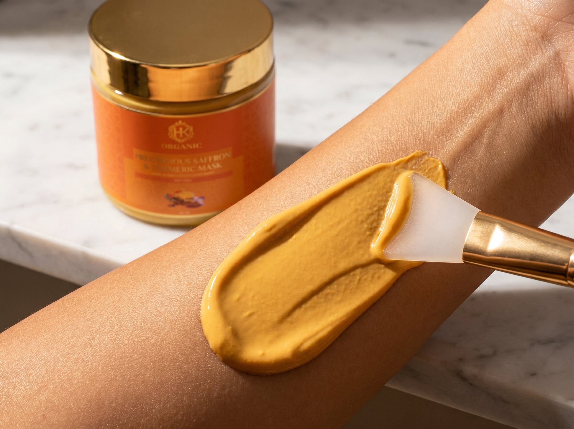 Gold-colored cosmetic product being applied to a person's arm with a brush, next to an orange jar on a marble surface.