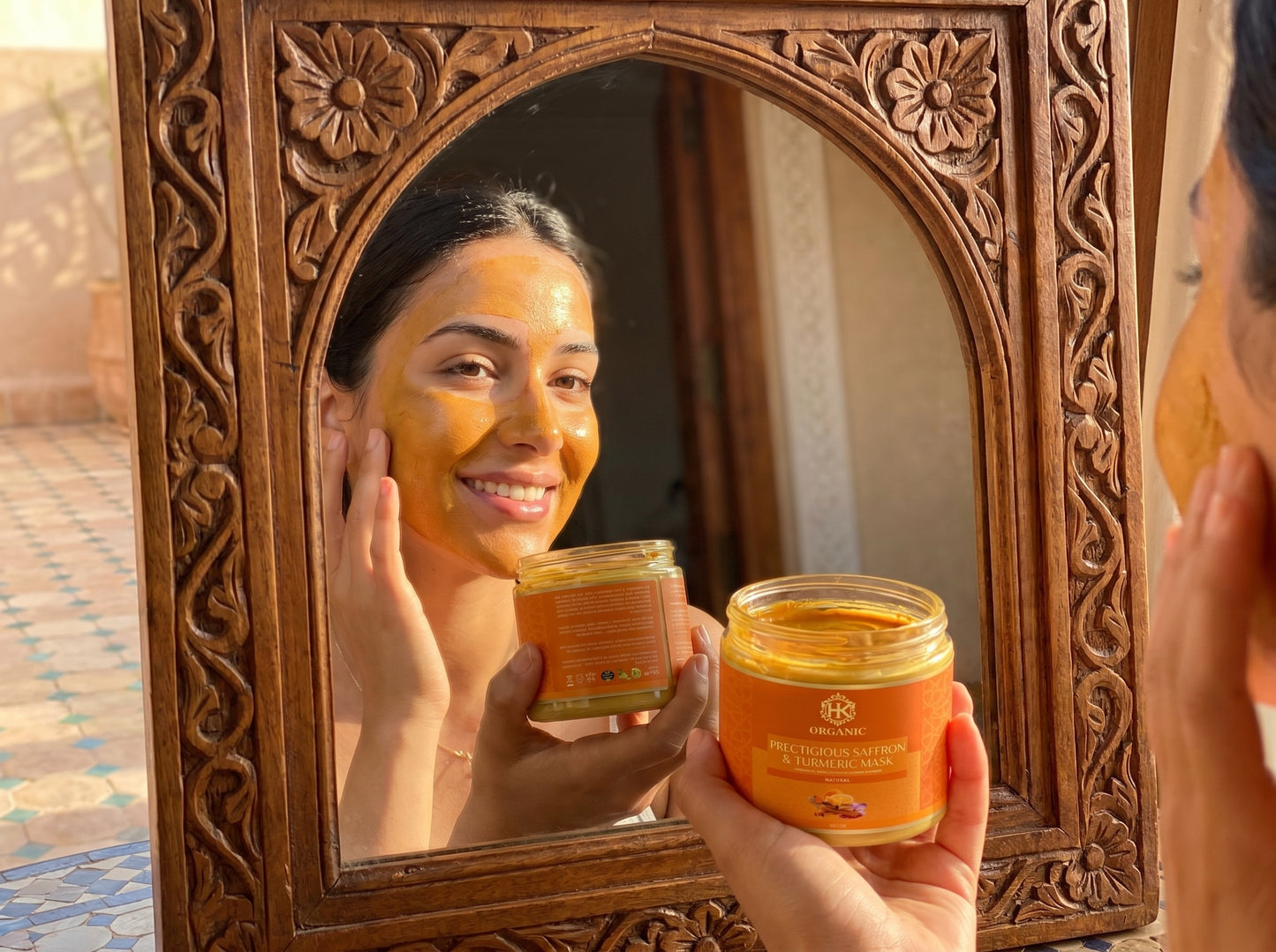 Person applying a facial mask in front of a decorative mirror, holding jars of the product.
