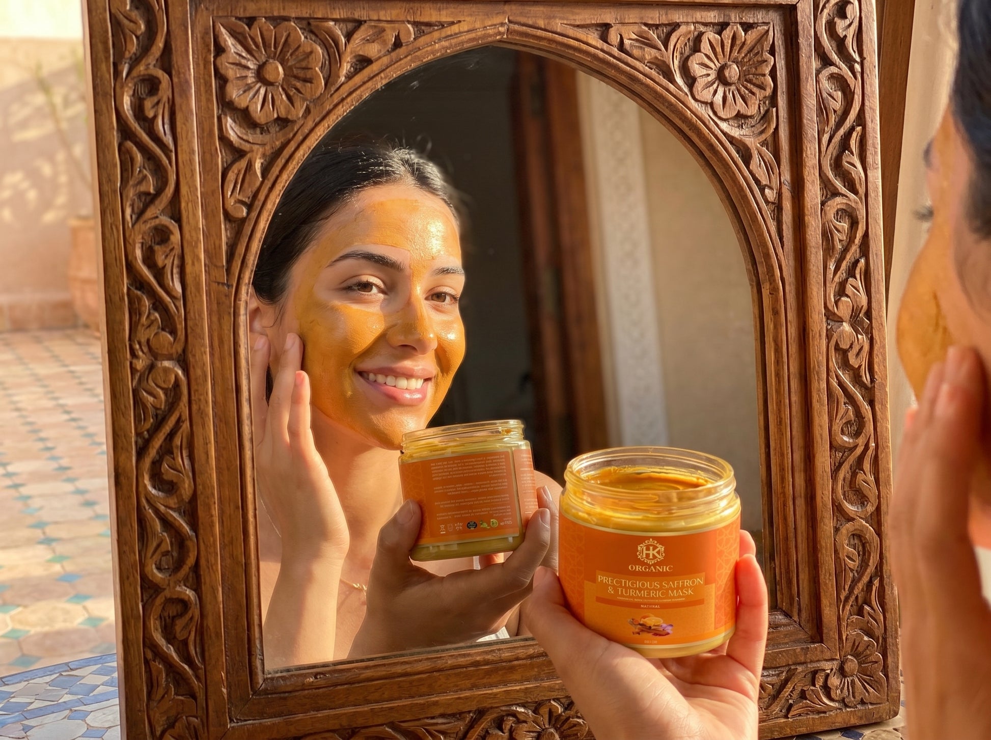 Person applying a facial mask in front of a decorative mirror, holding jars of the product.
