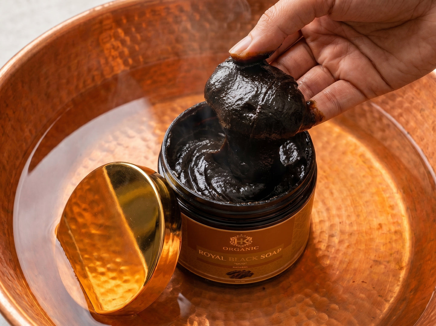 Hand scooping black soap from a jar labeled 'Royal Black Soap' on a copper tray.