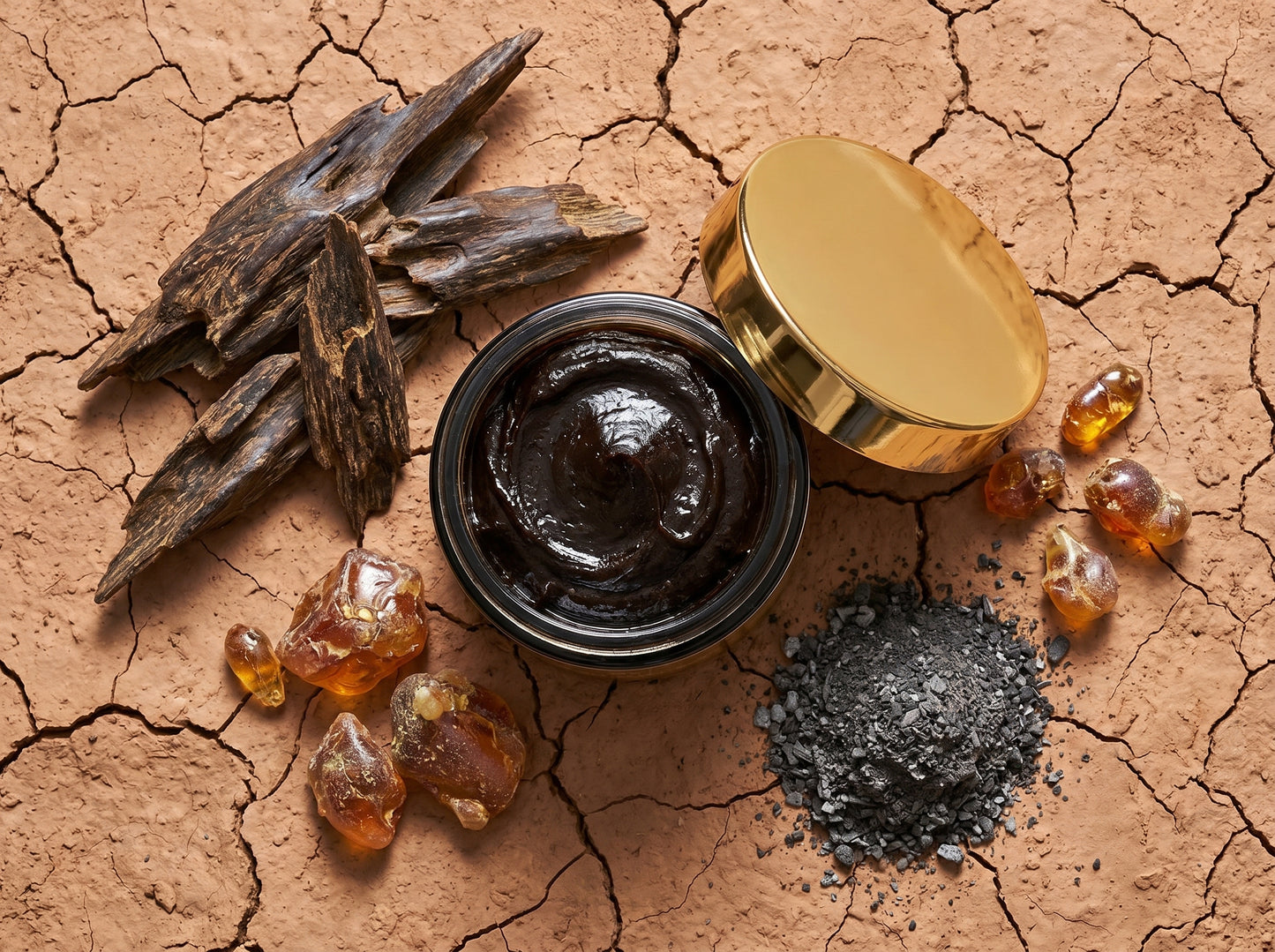 Open jar of dark brown paste with gold lid on a cracked brown surface with dried roots and amber-like substances.
