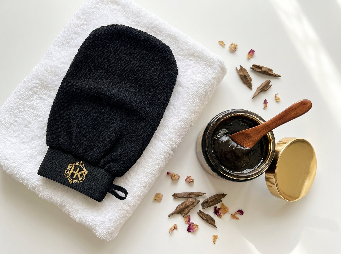 Black exfoliating glove with a brand logo on a white towel, next to a jar of dark paste and wooden spoon.