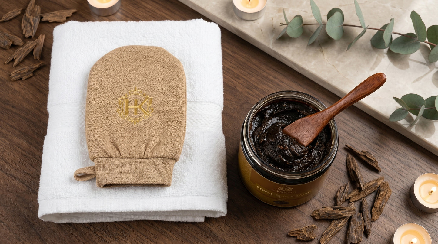 Brown exfoliating glove, jar of scrub, and wooden spoon on a wooden surface with towel and eucalyptus leaves.