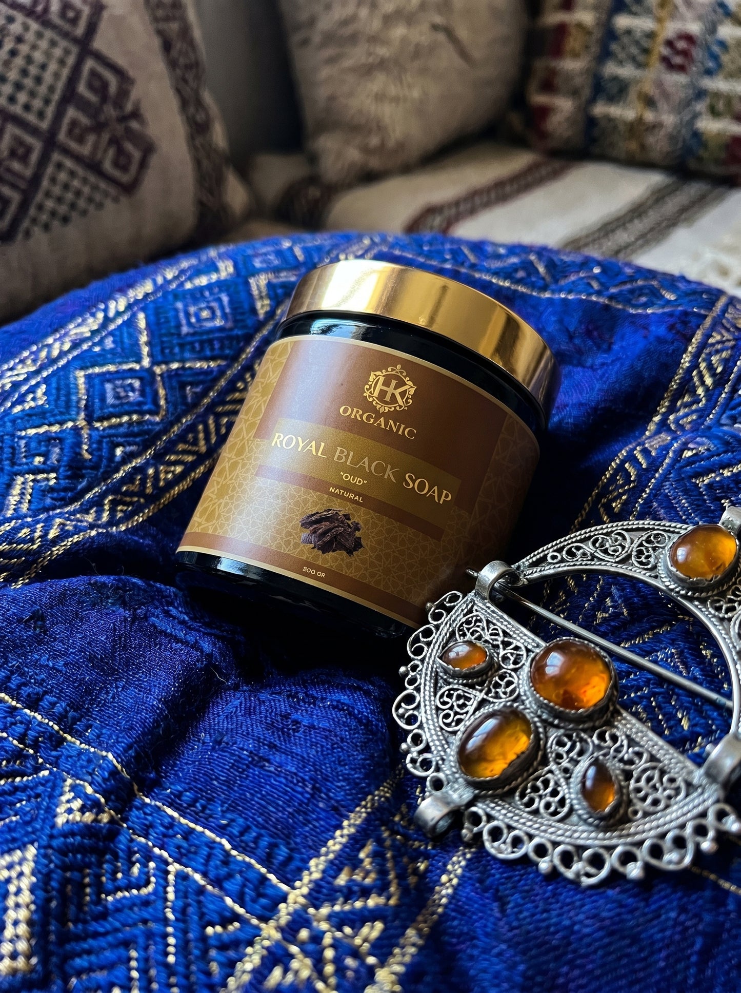 Jar of Royal Black Soap on a blue embroidered fabric with decorative earrings.