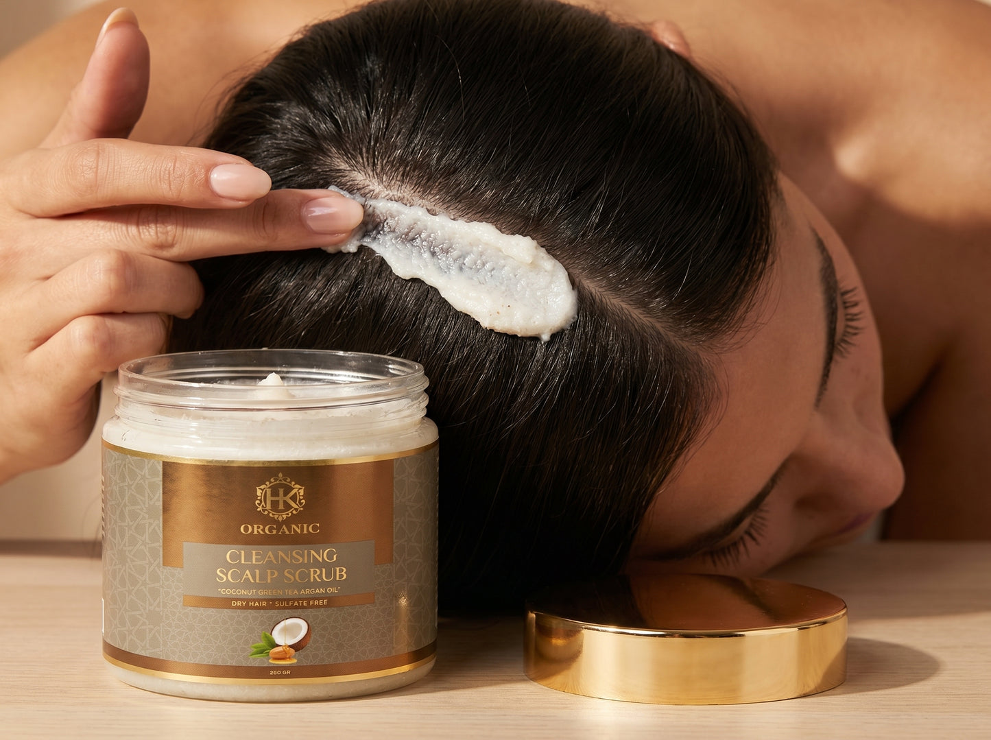 Person applying scalp scrub to their hair with a jar of the product on a wooden surface.