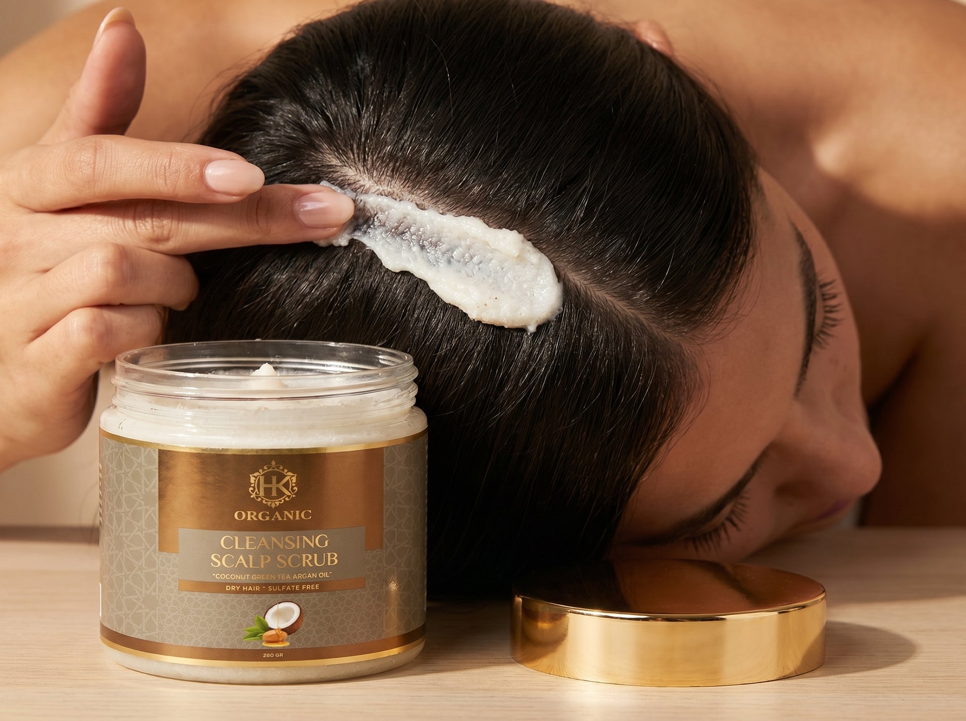Person applying scalp scrub to their hair with a jar of the product on a wooden surface.