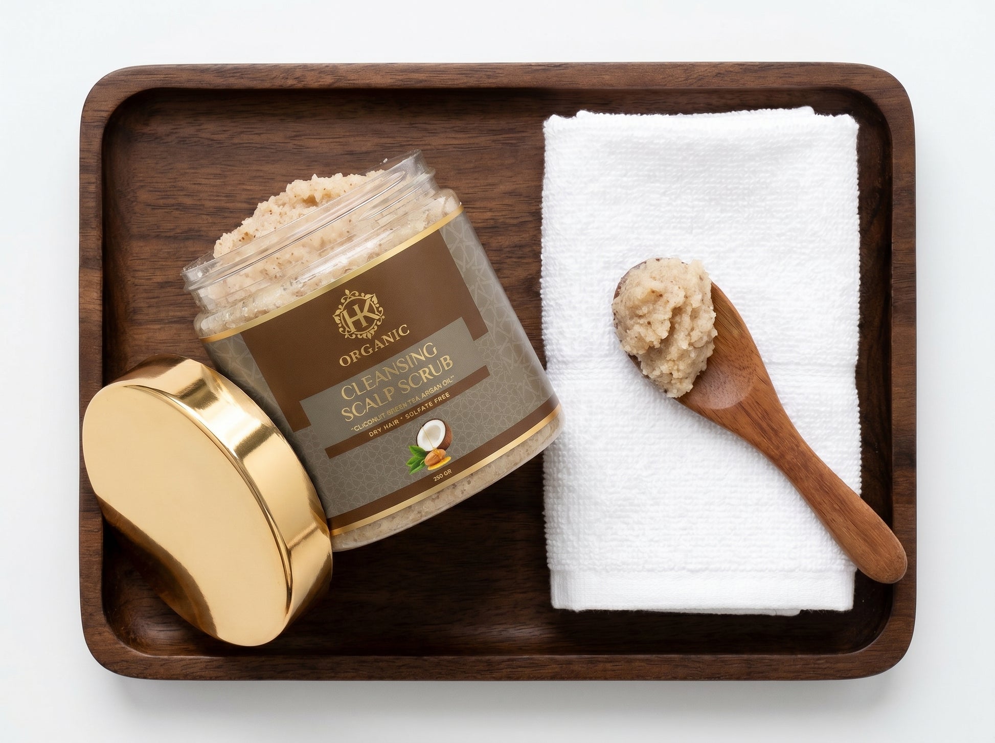 Jar of scalp scrub with a wooden spoonful on a white towel, all on a wooden tray.
