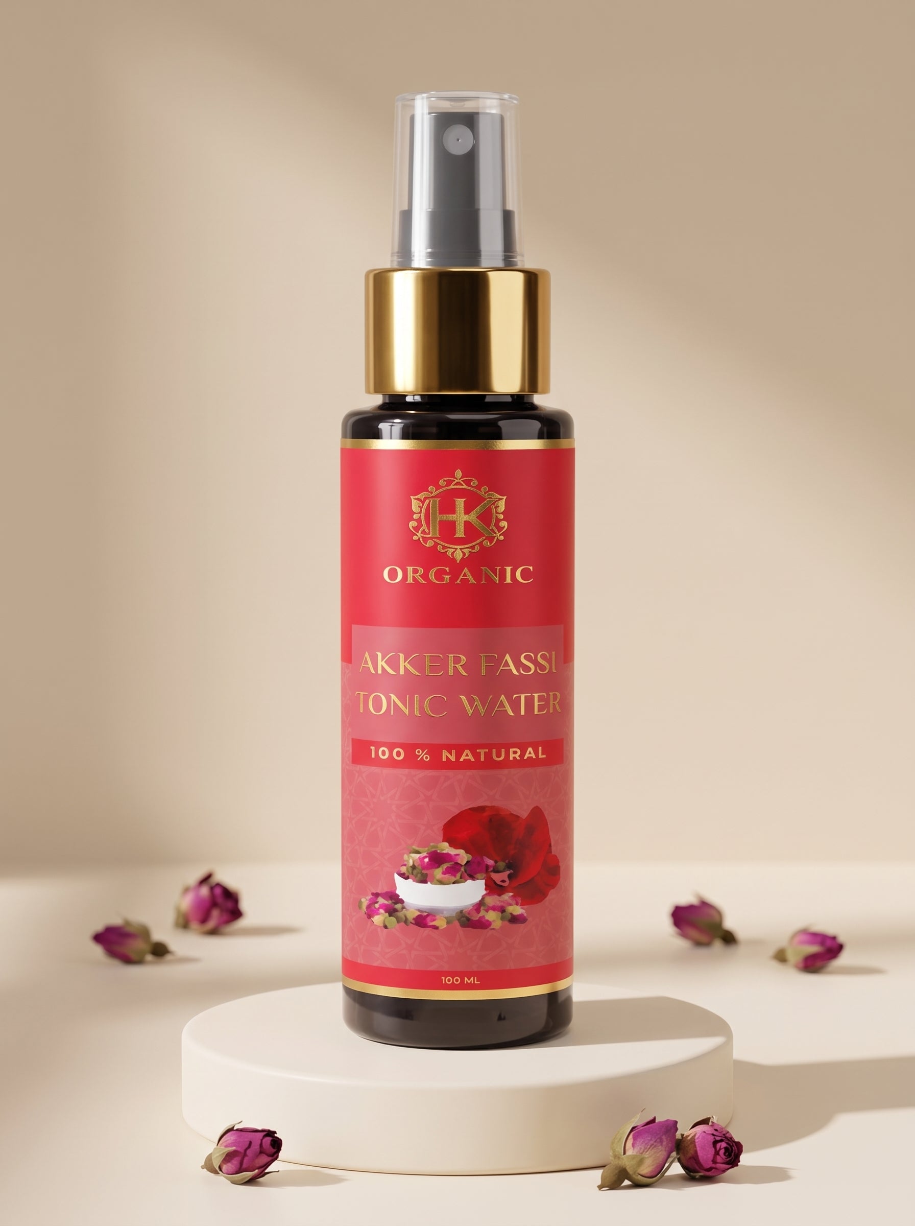 A bottle of Akker Fassa Organic Akker Fassa Ionic Water spray with a red label and gold pump, displayed on a white pedestal with scattered rose petals against a beige background.