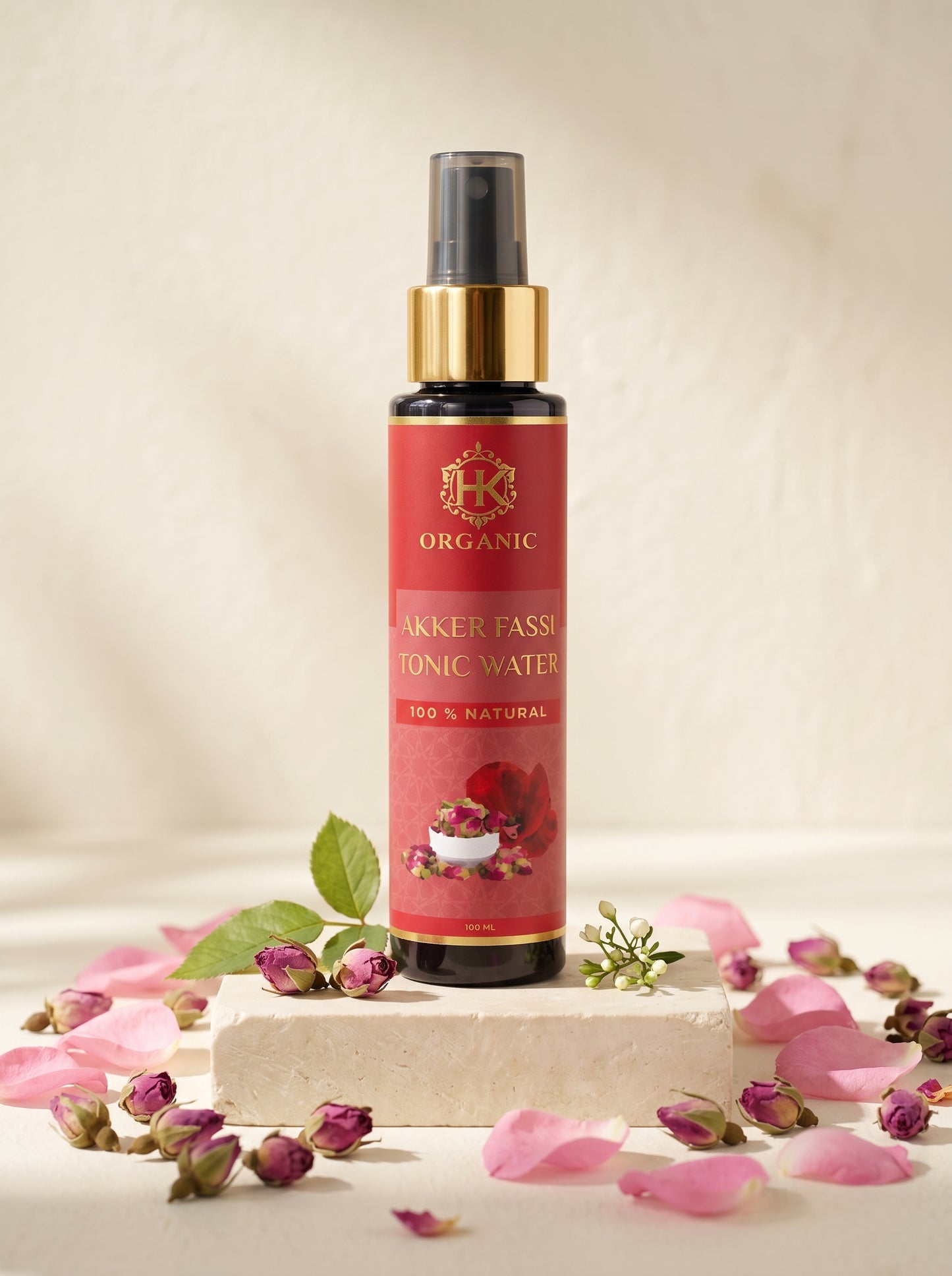 A bottle of Organic Akker Fassi Tonic Water with a red label and gold collar, displayed on a stone block surrounded by pink rose petals and buds.