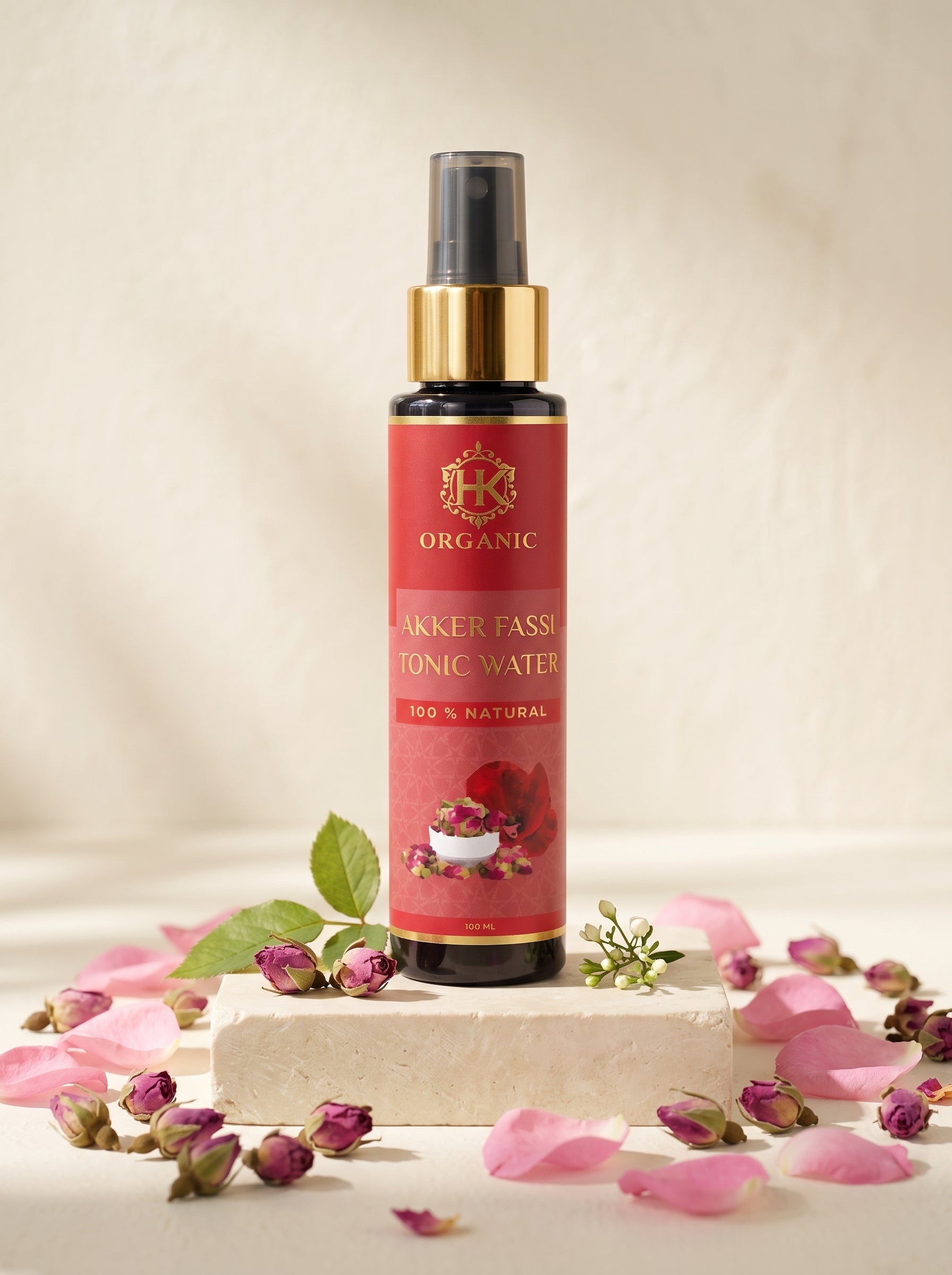 A bottle of Organic Akker Fassi Tonic Water with a red label and gold collar, displayed on a stone block surrounded by pink rose petals and buds.