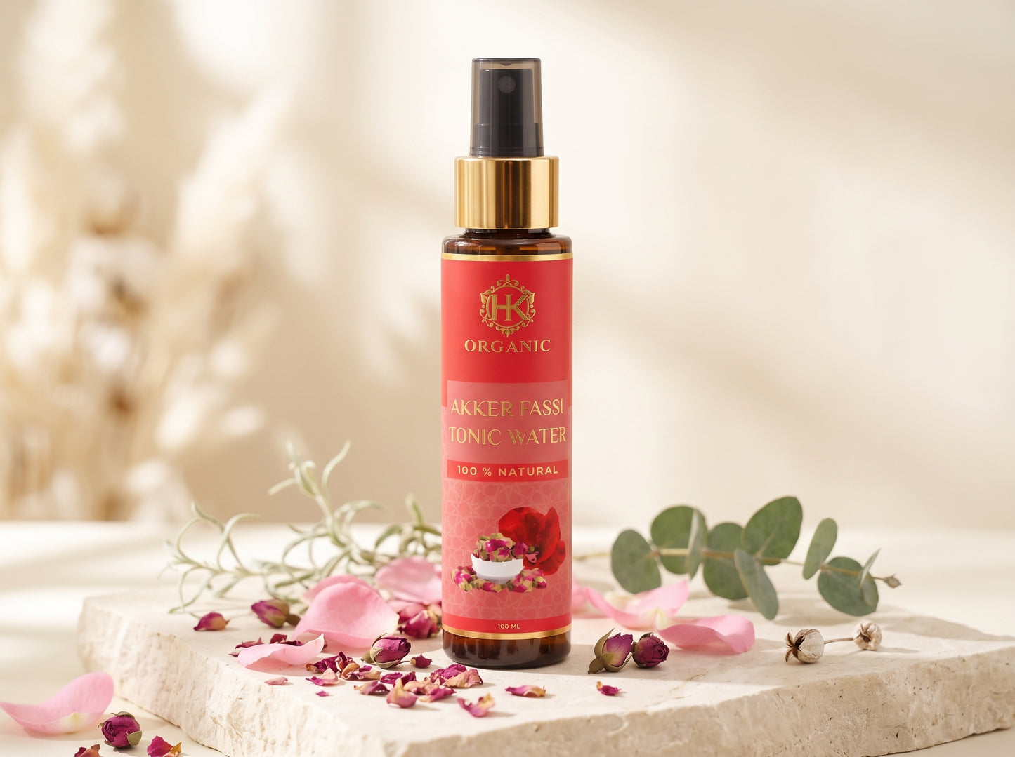 A bottle of Organic Sancker Pass Ionic Water spray, styled on a light stone slab with pink petals, dried rosebuds, and green eucalyptus leaves against a warm, soft-focus background.
