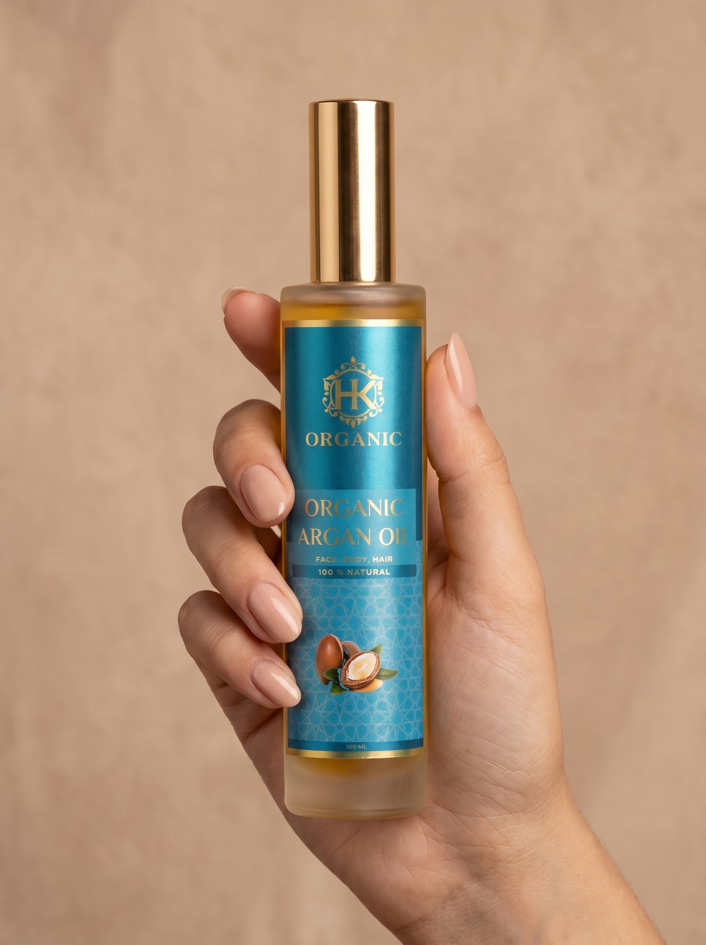 A hand holding a bottle of Organic Argan Oil against a warm beige textured background. The bottle has a gold cap and a blue label with gold lettering and a coconut illustration.