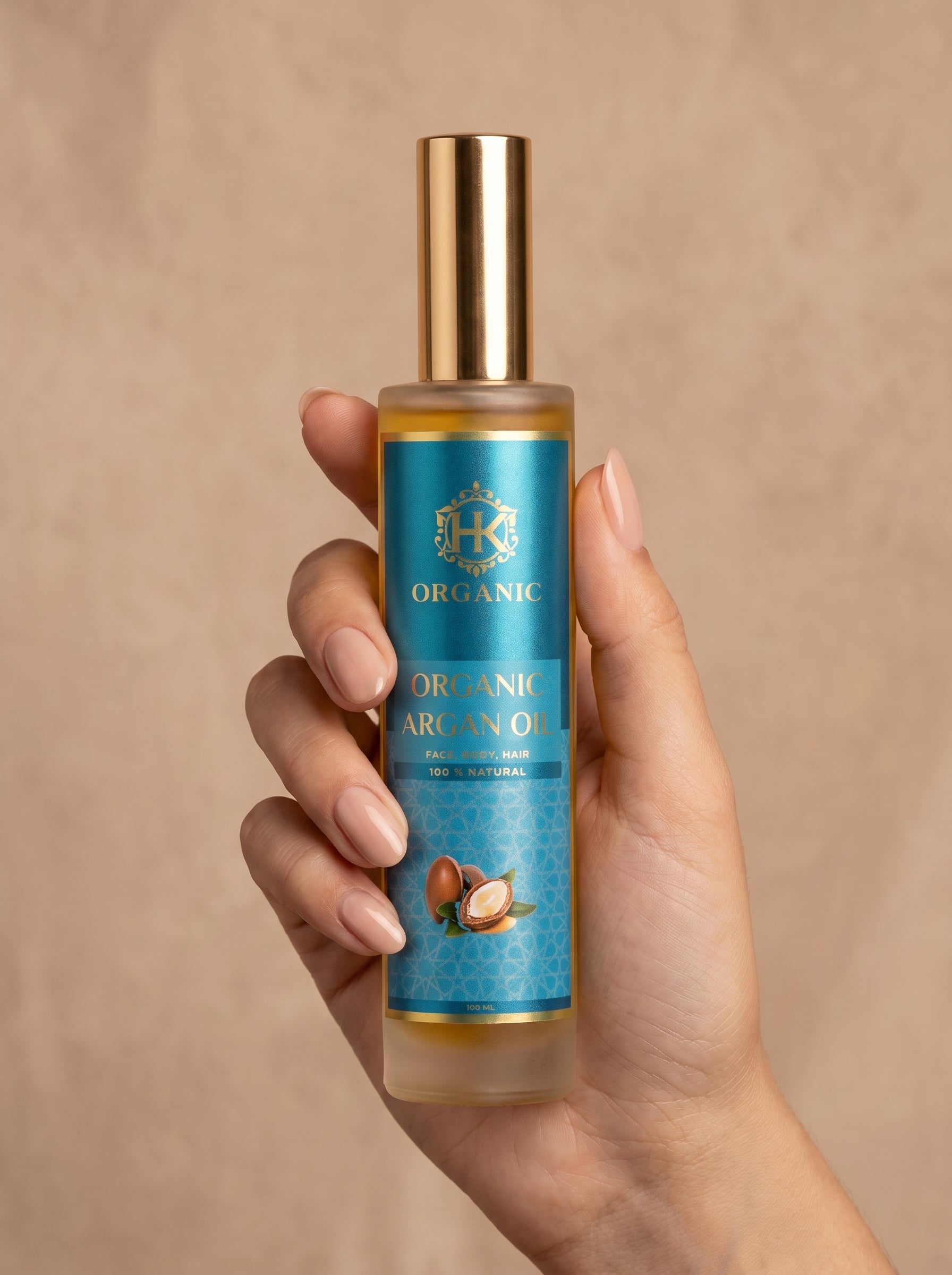 A hand holding a bottle of Organic Argan Oil against a warm beige textured background. The bottle has a gold cap and a blue label with gold lettering and a coconut illustration.