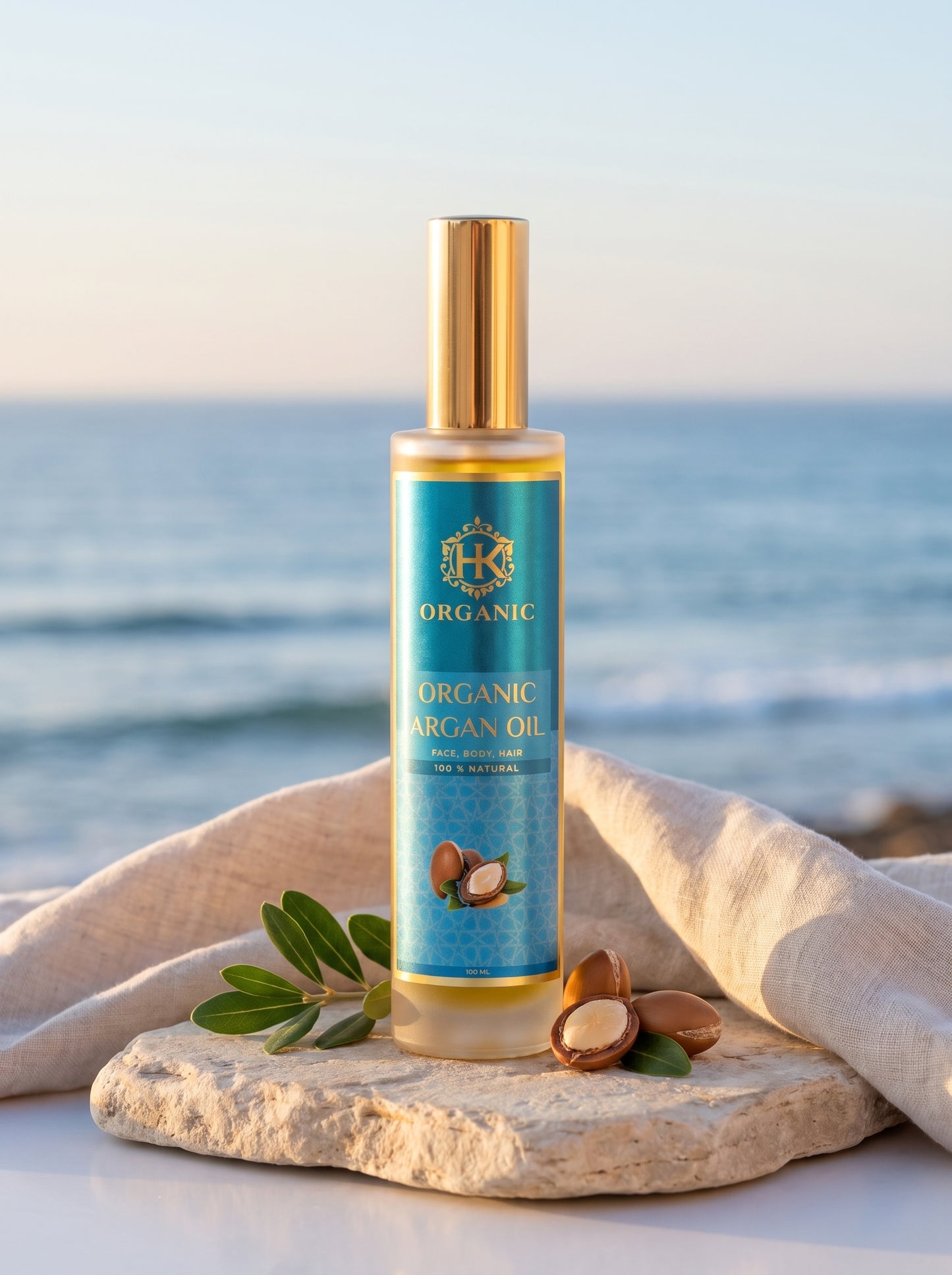 A bottle of Organic Argan Oil stands on a stone slab with a linen cloth and argan nuts, set against a blurred beach background at sunset.