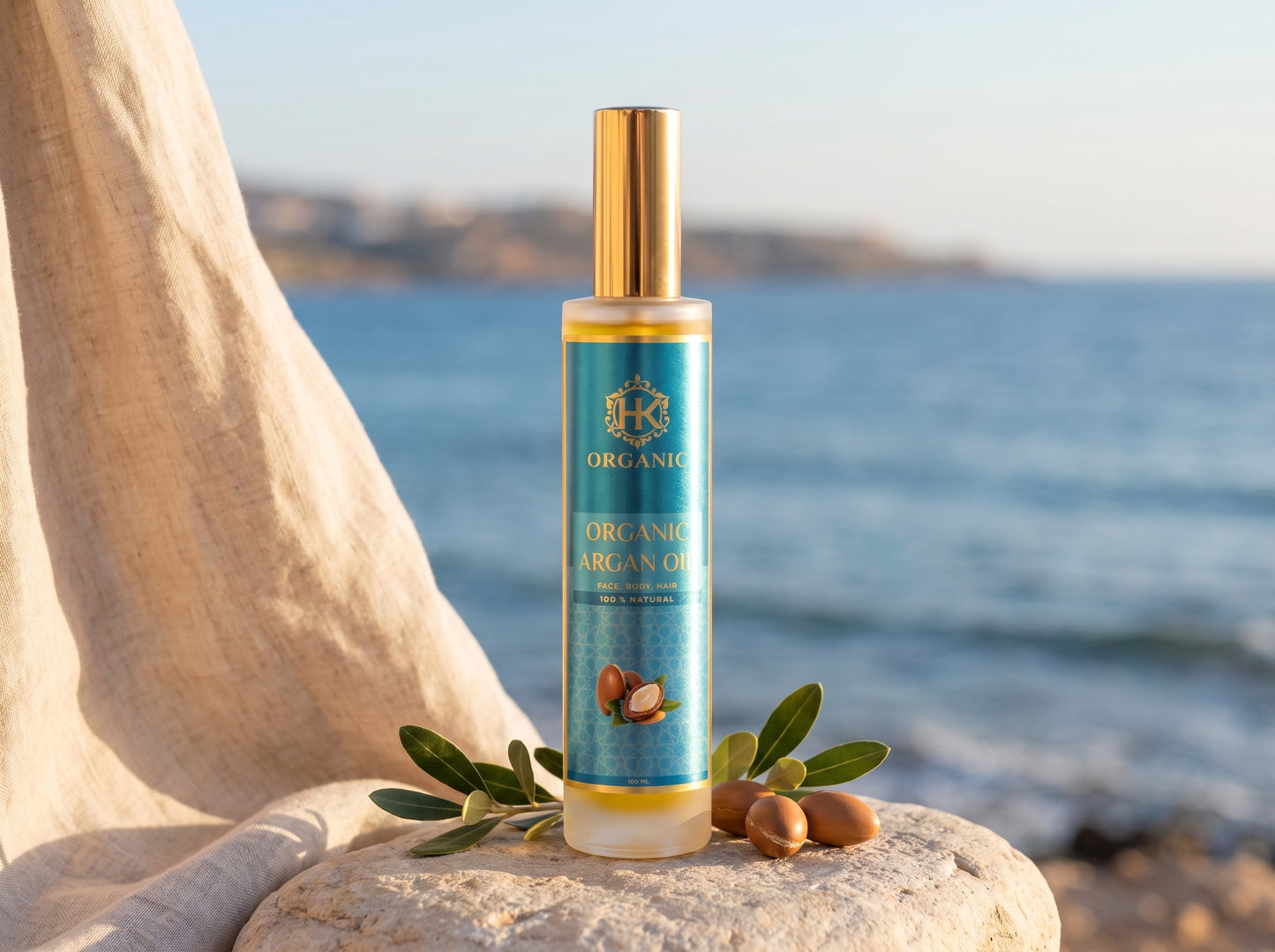 A bottle of Organic Argan Oil stands on a stone with argan nuts and green leaves, set against a blurred beach and sea background.