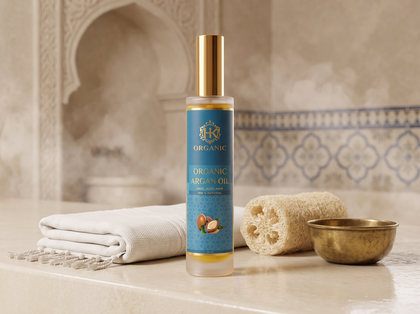 A bottle of Organic Argan Oil stands on a marble surface with a folded white towel, a natural sponge, and a small brass bowl against a warm, decorative tiled background.