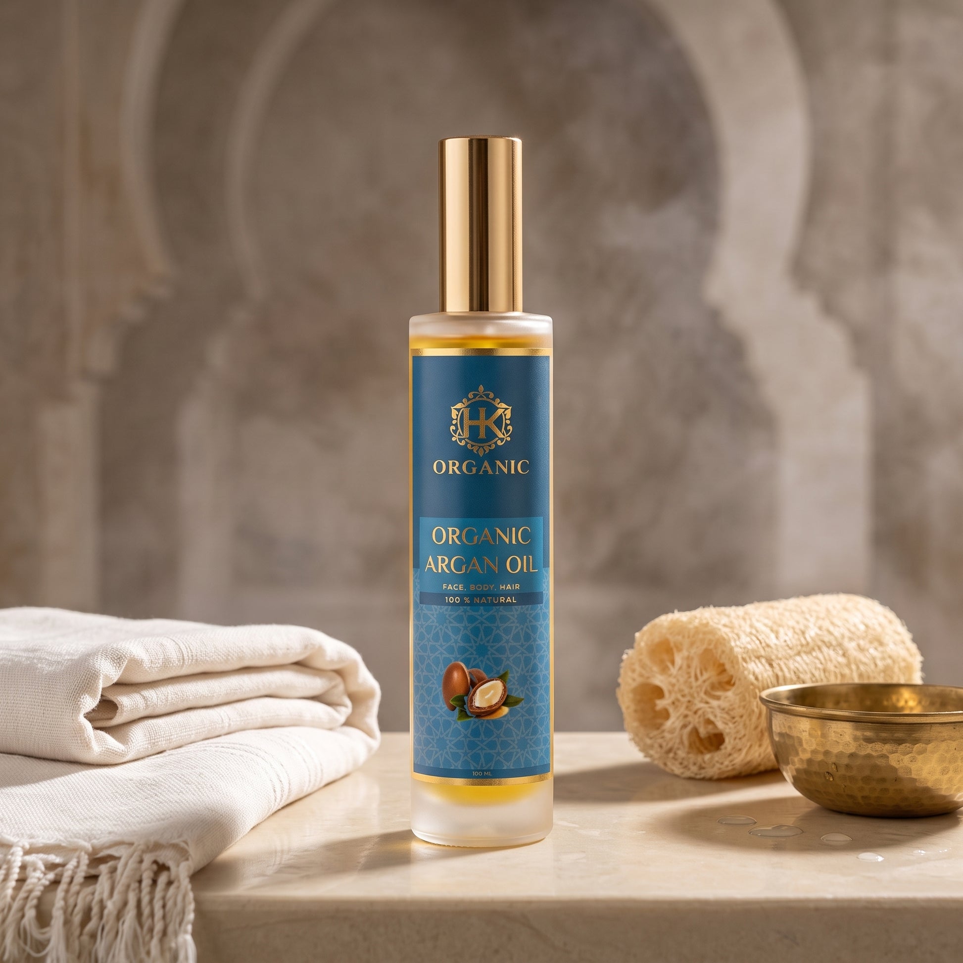 A bottle of Organic Argan Oil bath soak stands on a stone surface with a folded white towel, a natural loofah sponge, and a small brass bowl against a blurred stone arch background.